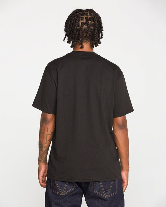 A person with short, twisted hair and a tattoo on their left arm is standing with their back to the camera, wearing the Volcom Men's Dual Euro Short Sleeve Tee in plain black and dark jeans against a plain white background.