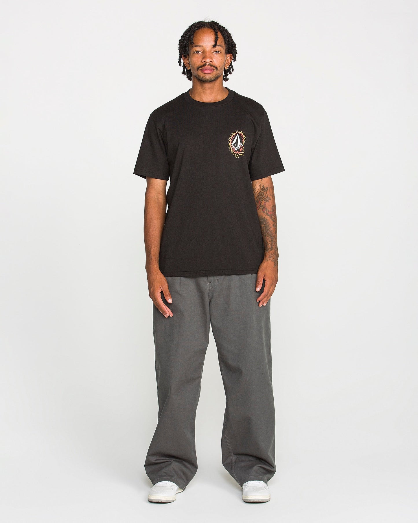 A person stands facing forward against a plain white background, wearing the Volcom Men's Ringafire Short Sleeve Tee in black—crafted from heavy-weight premium cotton with a small chest graphic—paired with gray wide-leg pants and white sneakers.
