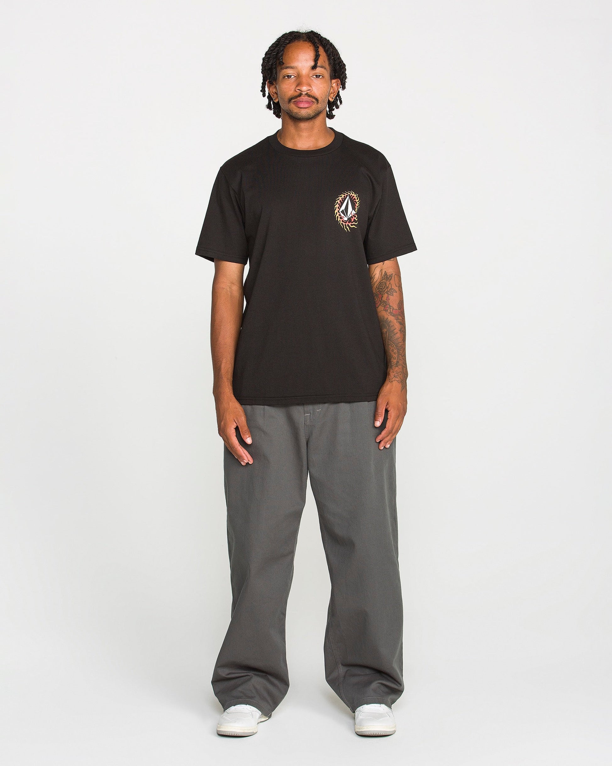 A person stands facing forward against a plain white background, wearing the Volcom Men's Ringafire Short Sleeve Tee in black—crafted from heavy-weight premium cotton with a small chest graphic—paired with gray wide-leg pants and white sneakers.