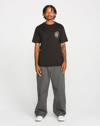 A person stands facing forward against a plain white background, wearing the Volcom Men's Ringafire Short Sleeve Tee in black—crafted from heavy-weight premium cotton with a small chest graphic—paired with gray wide-leg pants and white sneakers.