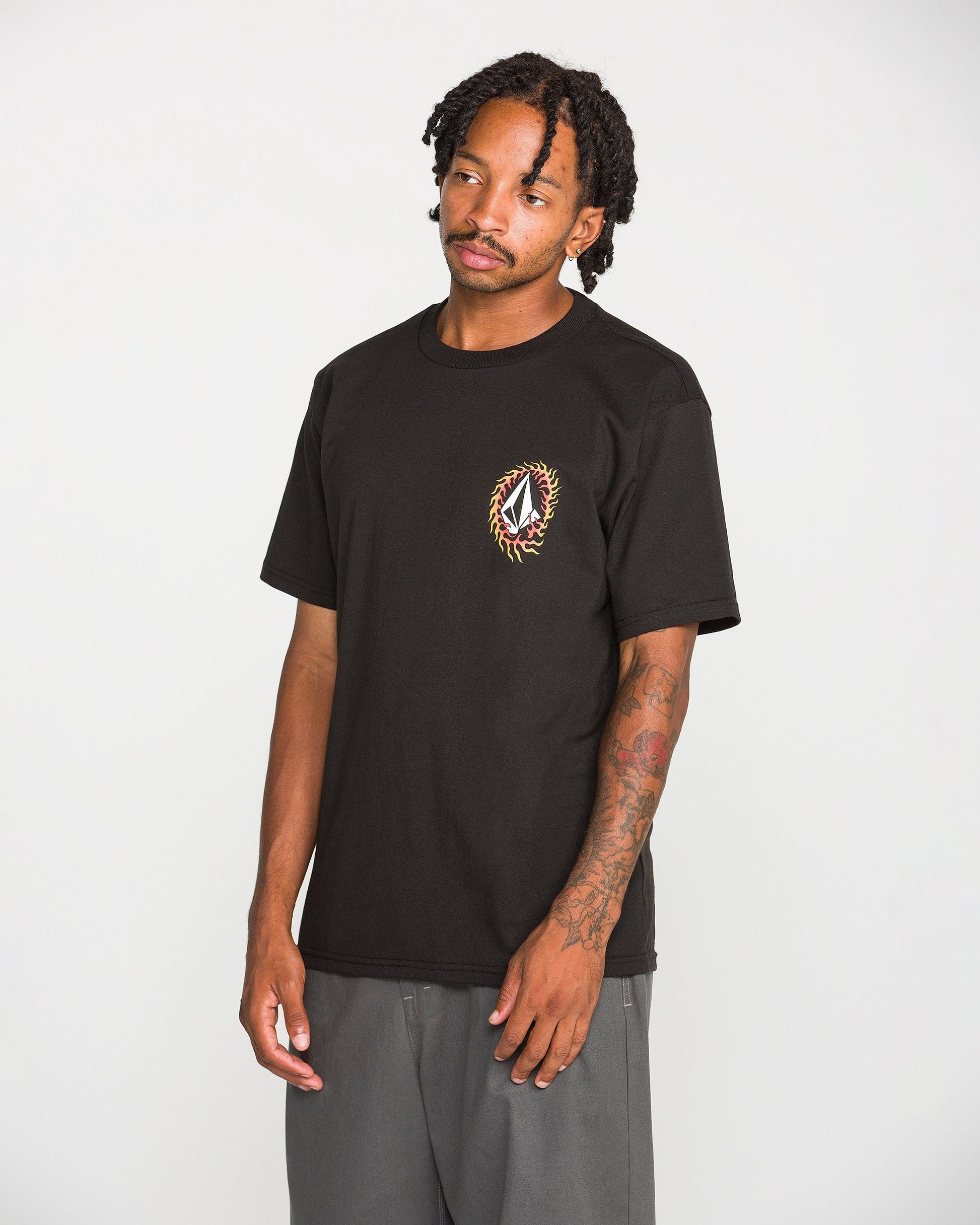 A man with tattoos and dreadlocks stands against a plain white background, wearing the Volcom Men's Ringafire Short Sleeve Tee—a classic fit black t-shirt made from heavy-weight premium cotton, featuring a small, colorful embroidered design on the left chest—paired with gray pants.