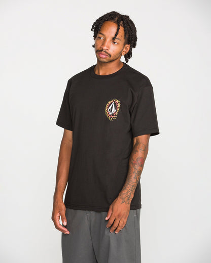 A man with tattoos and dreadlocks stands against a plain white background, wearing the Volcom Men's Ringafire Short Sleeve Tee—a classic fit black t-shirt made from heavy-weight premium cotton, featuring a small, colorful embroidered design on the left chest—paired with gray pants.