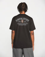 A person with short dreadlocks is seen from behind, wearing a black Volcom Men's Ringafire Short Sleeve Tee made of heavy-weight premium cotton, featuring white graphic text that reads “KINGDOM SPORTING ESTD 1991 ROAD-TESTED WORLDWIDE.” They are also wearing gray shorts.