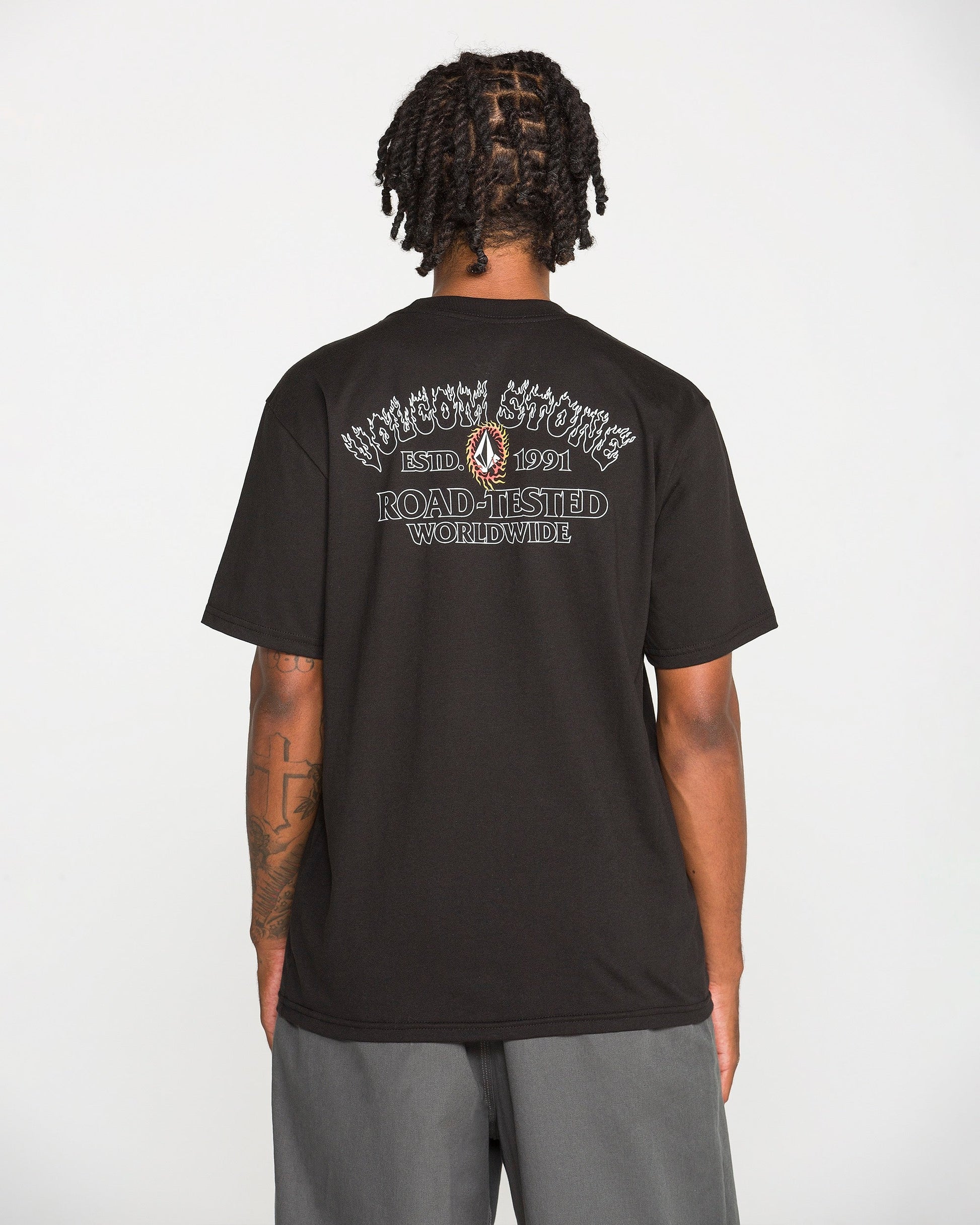 A person with short dreadlocks is seen from behind, wearing a black Volcom Men's Ringafire Short Sleeve Tee made of heavy-weight premium cotton, featuring white graphic text that reads “KINGDOM SPORTING ESTD 1991 ROAD-TESTED WORLDWIDE.” They are also wearing gray shorts.