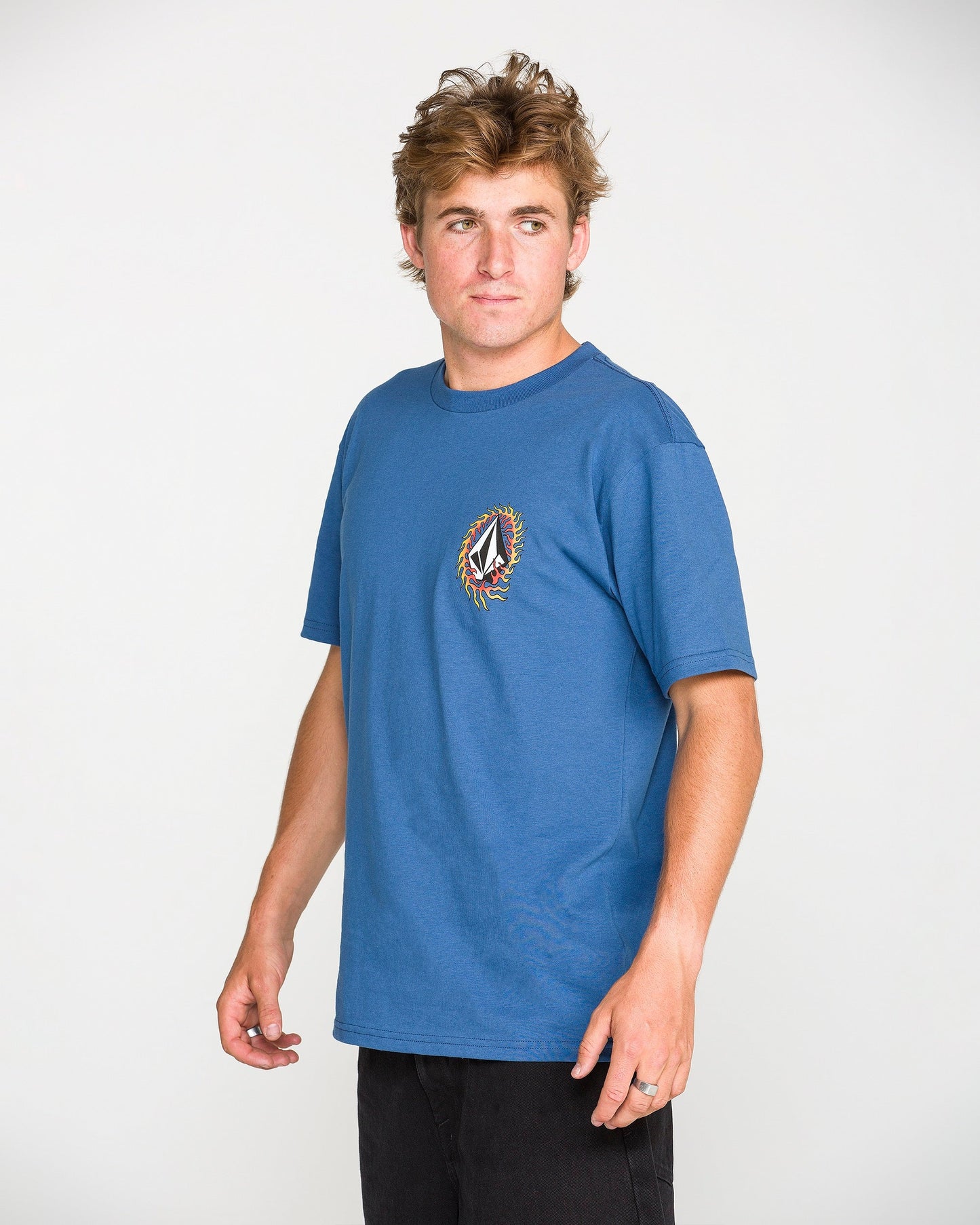 A person with tousled hair, wearing the Volcom Men's Ringafire Short Sleeve Tee in blue—crafted from premium ring spun cotton and featuring a colorful chest logo—stands turned slightly to the side against a plain white background.