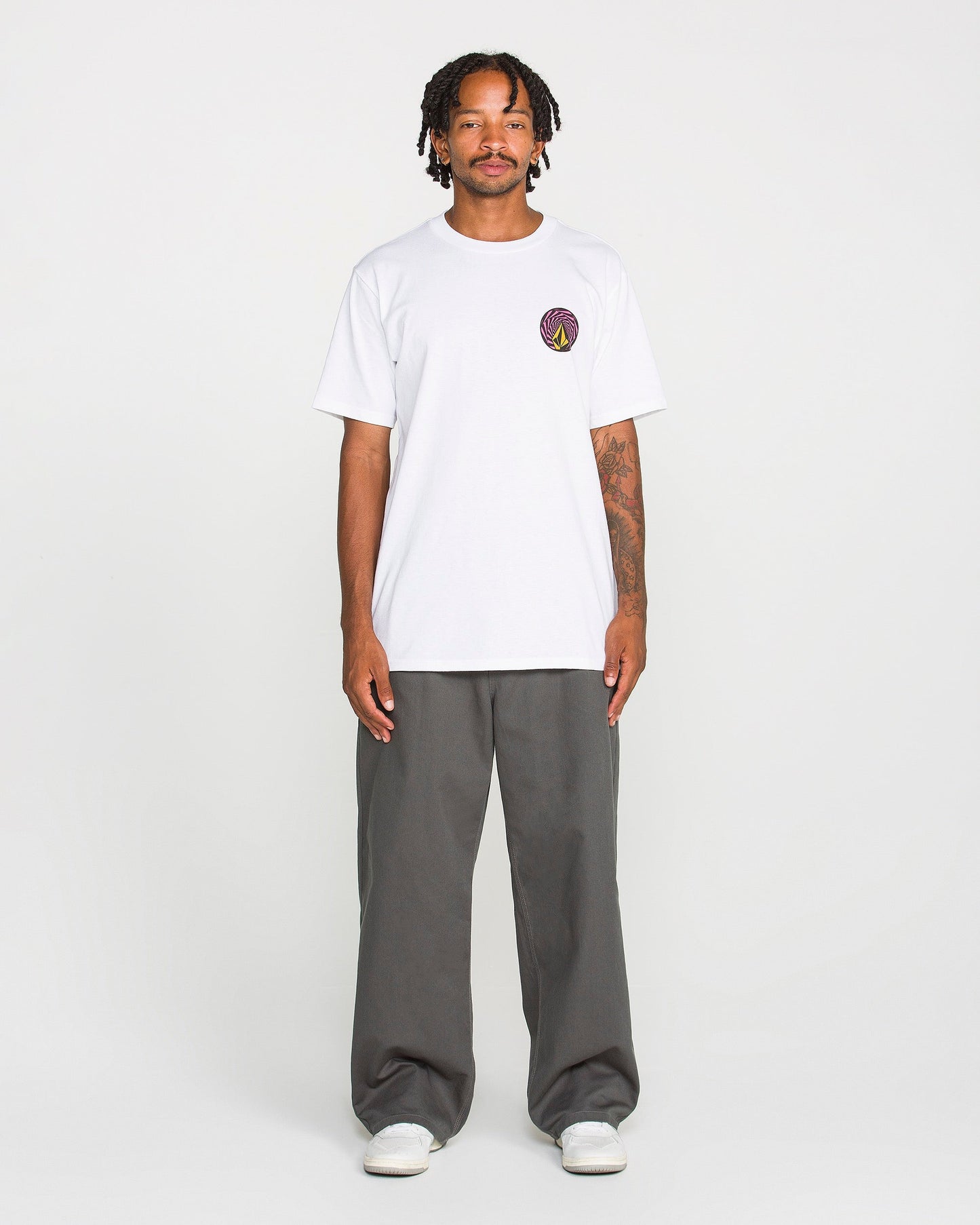 A person stands facing forward, wearing the Volcom Men's Dirty Work Short Sleeve Tee—a classic fit made from heavyweight cotton with a small soft-hand screen print logo on the left chest—paired with gray pants and white shoes, against a plain white background.