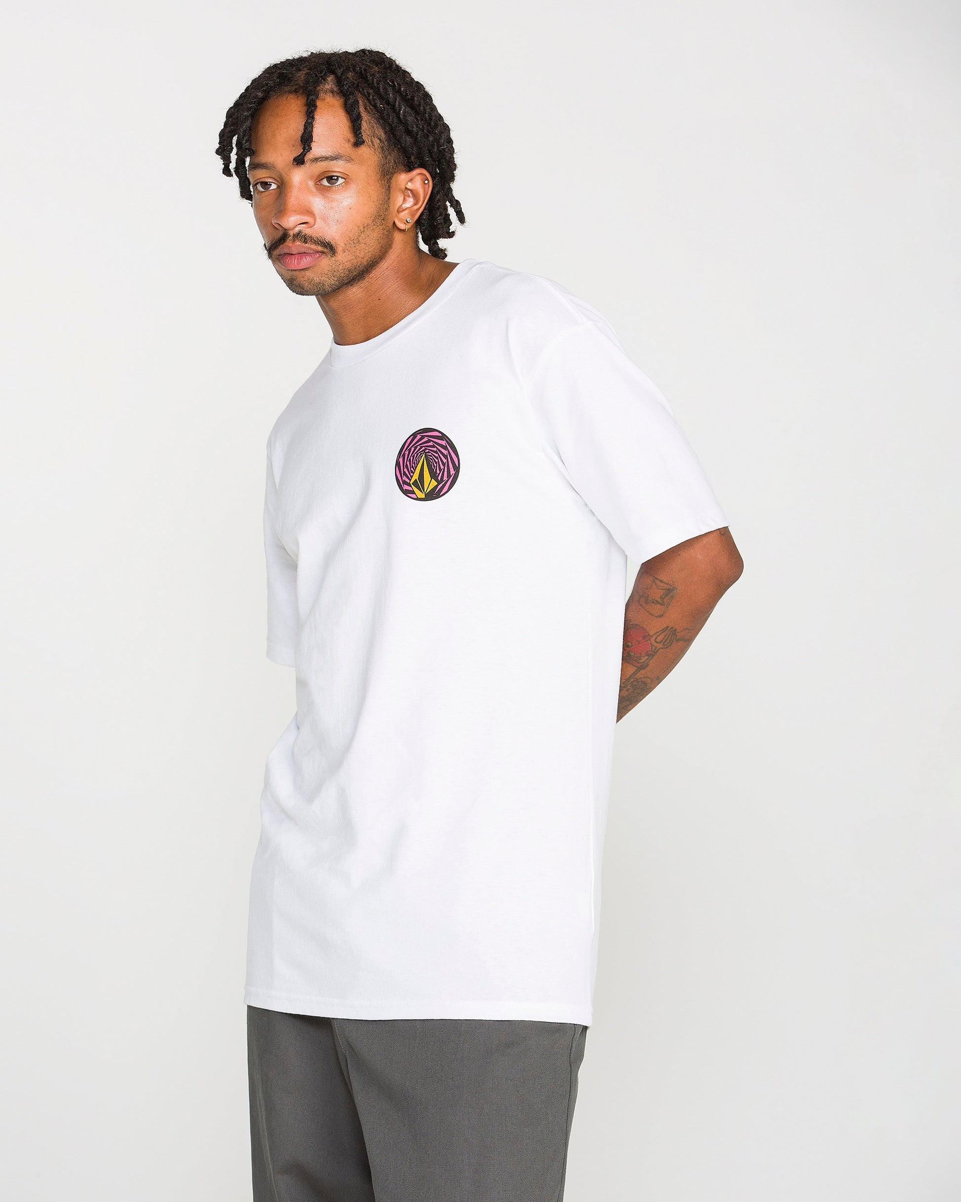 A man with short dreadlocks wears the Volcom Men's Dirty Work Short Sleeve Tee, crafted from heavyweight cotton and featuring a colorful circular logo on the chest in a soft-hand screen print. He pairs it with gray pants while standing against a plain, light background.