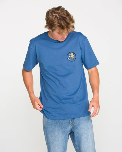 A person dressed in jeans and a blue, classic fit Volcom Men's Stoneyguy Short Sleeve Tee looks down while holding the shirt’s hem. Made from premium cotton, the t-shirt features a small, round soft-hand screen print graphic on the left chest. The background is plain white.