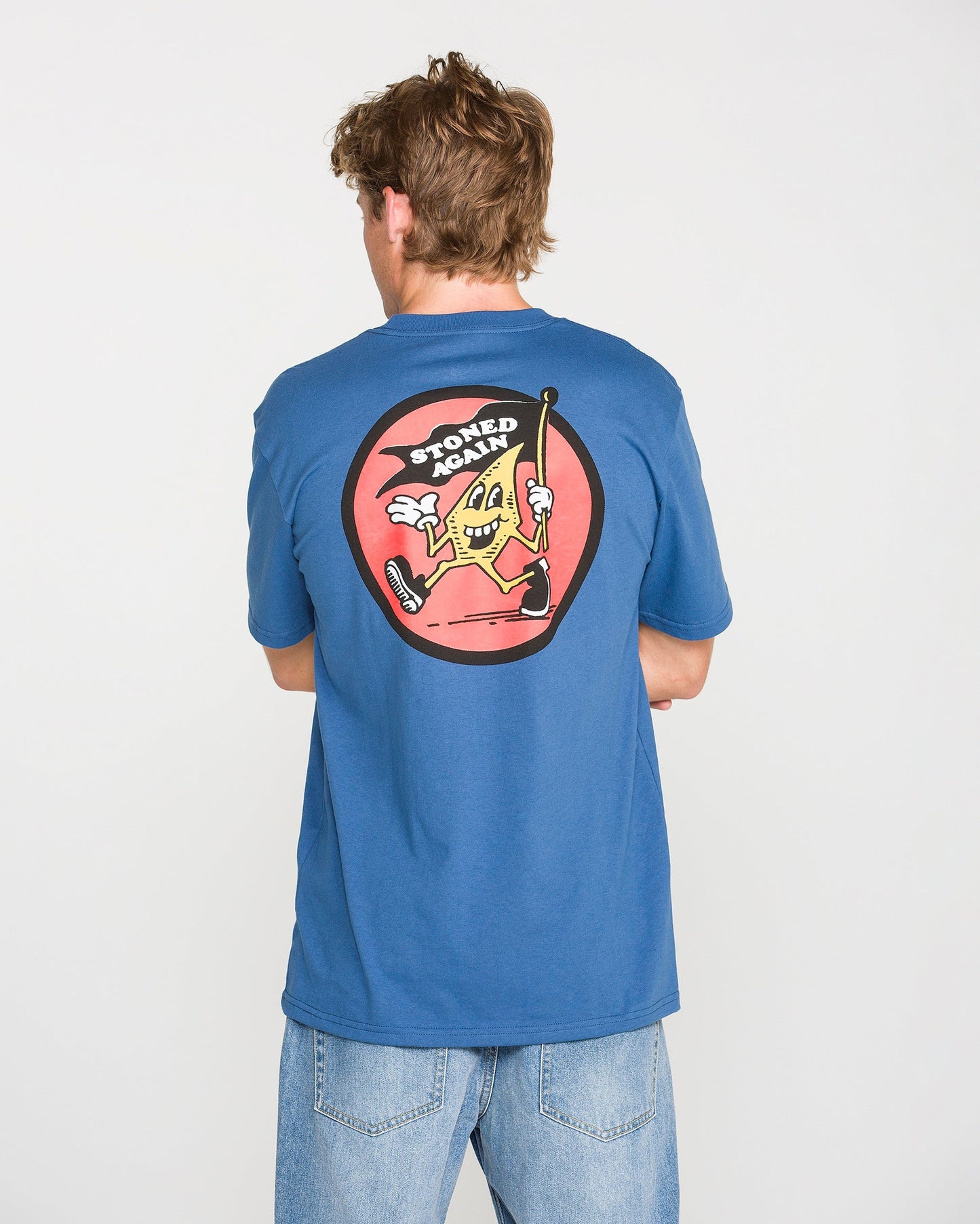 A person wearing the Volcom Men's Stoneyguy Short Sleeve Tee in blue and light jeans is seen from behind. The shirt features a soft-hand screen print of a cartoon slice of pizza holding a cane, with the text "STONED AGAIN" above it in a red circle.