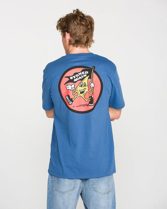 A person wearing the Volcom Men's Stoneyguy Short Sleeve Tee in blue and light jeans is seen from behind. The shirt features a soft-hand screen print of a cartoon slice of pizza holding a cane, with the text "STONED AGAIN" above it in a red circle.