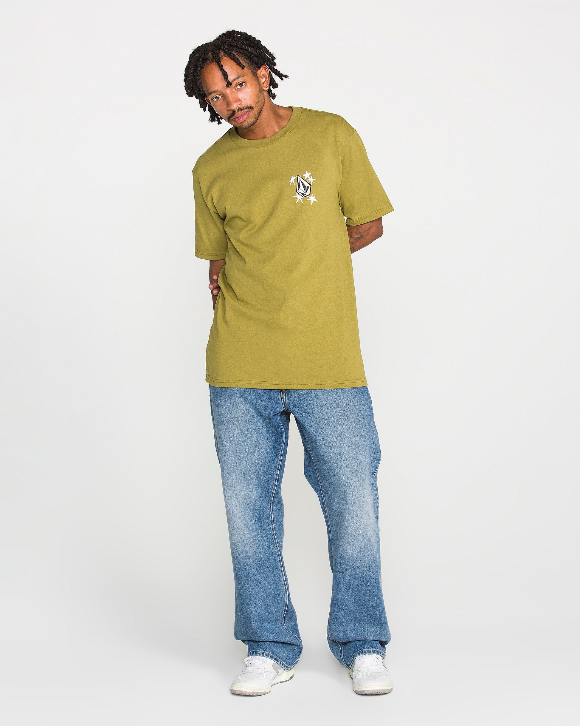 A person wearing the Volcom Men's Alive We Ride Short Sleeve Tee in yellow-green, featuring a soft-hand screen print, stands against a plain white background looking at the camera with their hands behind their back. The tee is crafted from heavyweight premium cotton for lasting comfort and style, and is paired with blue jeans and white sneakers.