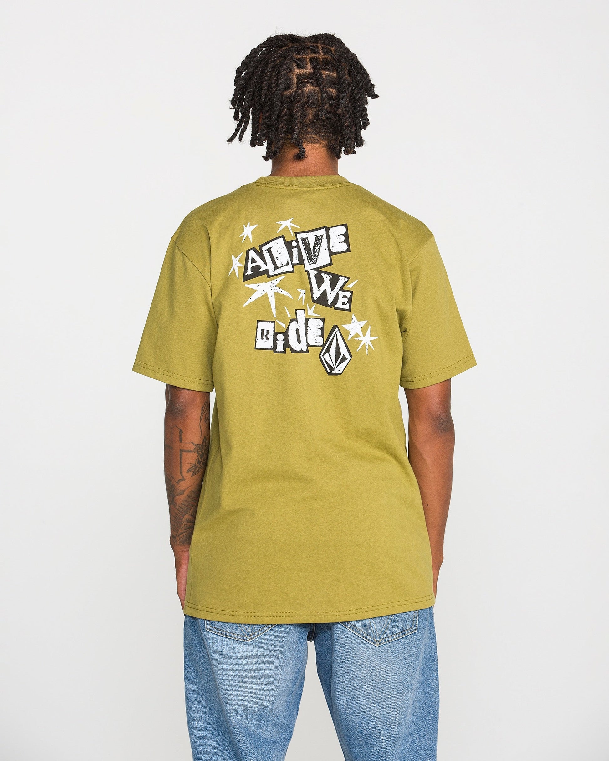 A person with short dreadlocks is shown from the back wearing the Volcom Men's Alive We Ride Short Sleeve Tee in olive green paired with blue jeans. This heavyweight premium cotton shirt features a soft-hand screen-printed graphic of shapes, a logo, and text that reads "ALIVE WE RIDE.