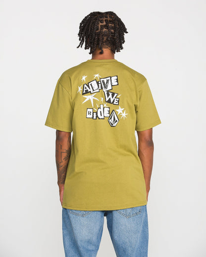A person with short dreadlocks is shown from the back wearing the Volcom Men's Alive We Ride Short Sleeve Tee in olive green paired with blue jeans. This heavyweight premium cotton shirt features a soft-hand screen-printed graphic of shapes, a logo, and text that reads "ALIVE WE RIDE.