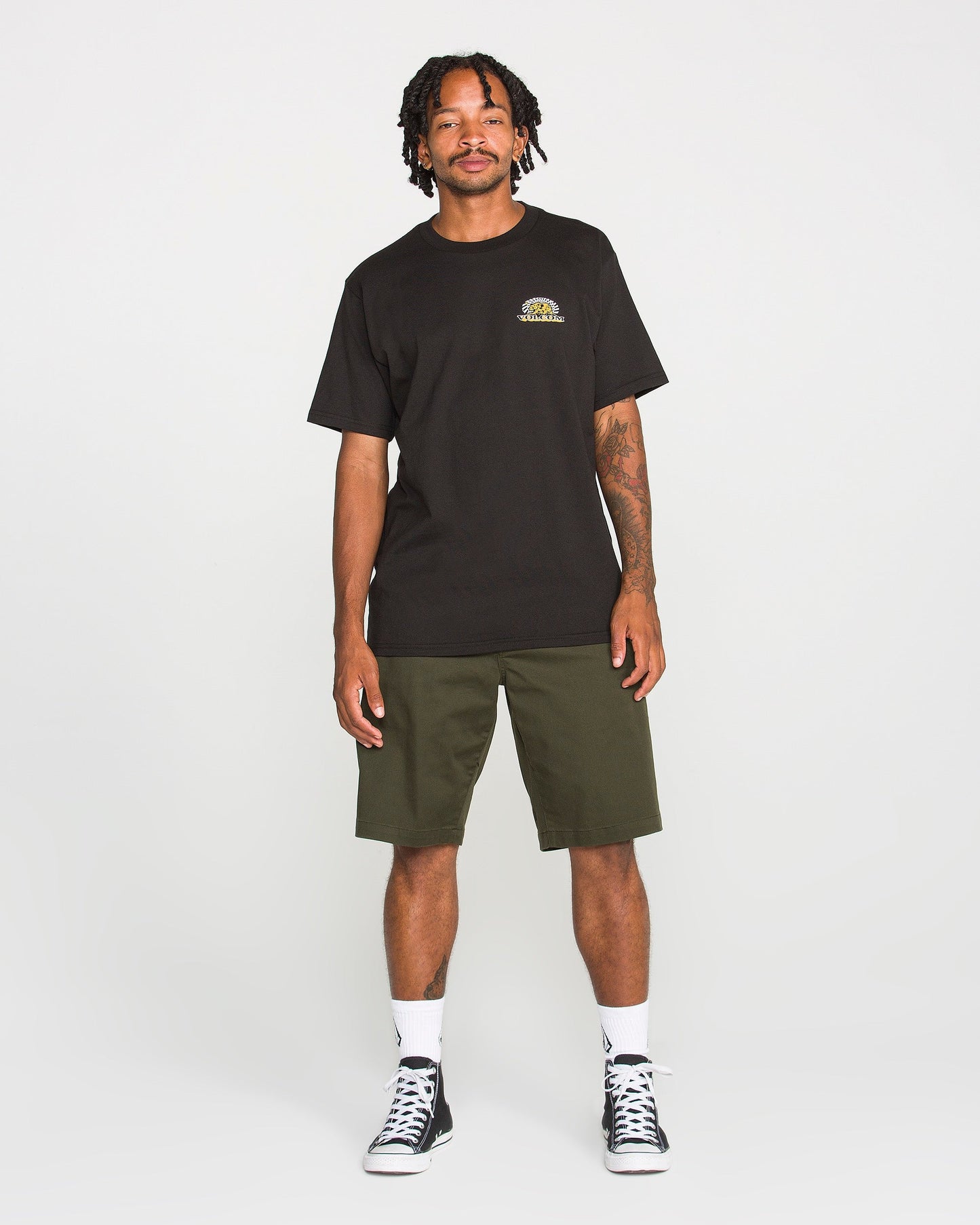 A man stands smiling in a Volcom Men's Golden Bear Short Sleeve Tee crafted from heavyweight cotton, paired with olive green shorts, white socks, and black sneakers against a plain white background.