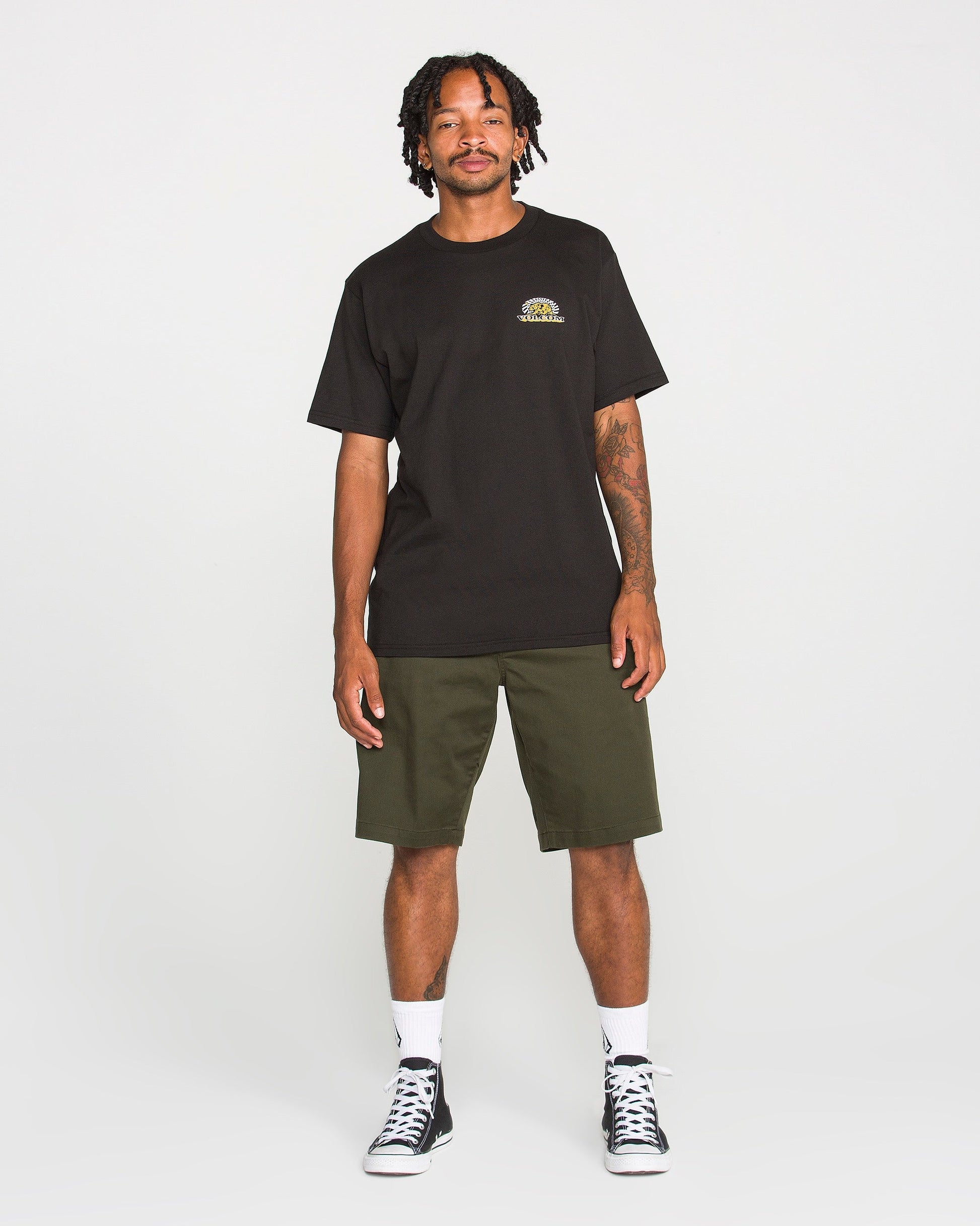 A man stands smiling in a Volcom Men's Golden Bear Short Sleeve Tee crafted from heavyweight cotton, paired with olive green shorts, white socks, and black sneakers against a plain white background.