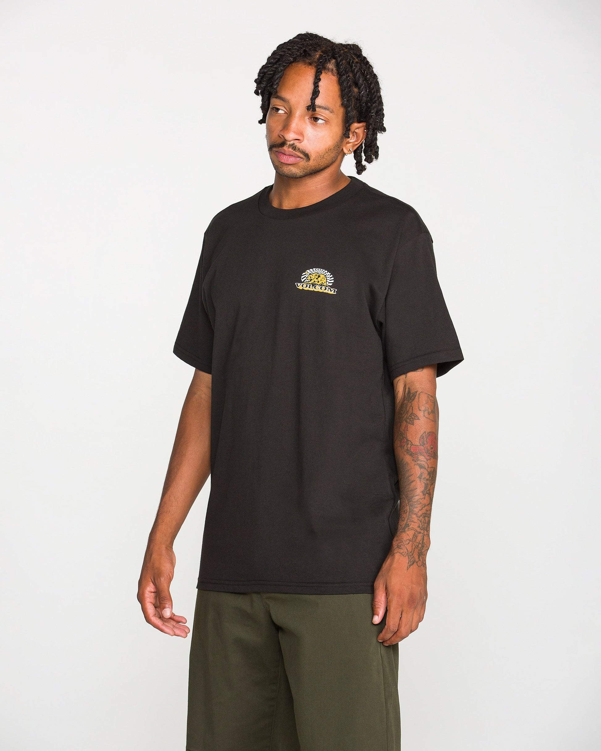A man wearing the Volcom Men's Golden Bear Short Sleeve Tee—a black heavyweight cotton T-shirt featuring a small round yellow Volcom Stone logo on the left chest—and olive green pants stands against a plain white background.