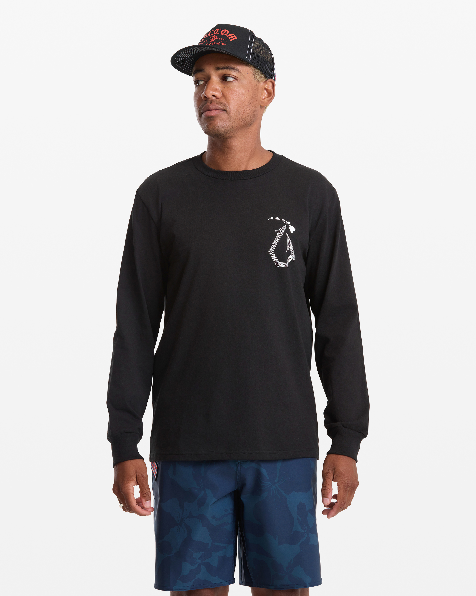 A man wears the Volcom Men's Makau Long Sleeve Tee in black, featuring a small white chest logo, paired with blue patterned shorts and a black and red cap, standing against a plain white background.