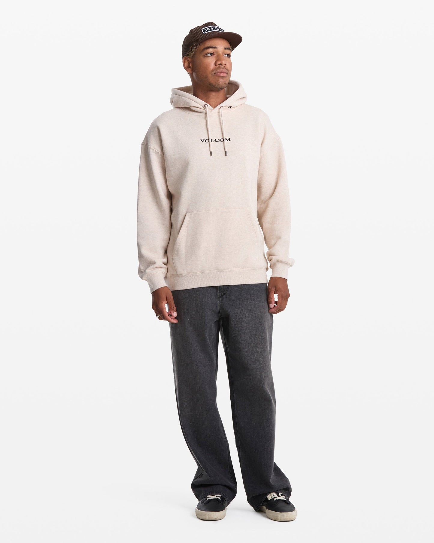 A man wears a beige Men's Volcom Stone Pullover by Volcom, paired with dark gray jeans, black sneakers, and a brown cap as he stands against a plain white background.