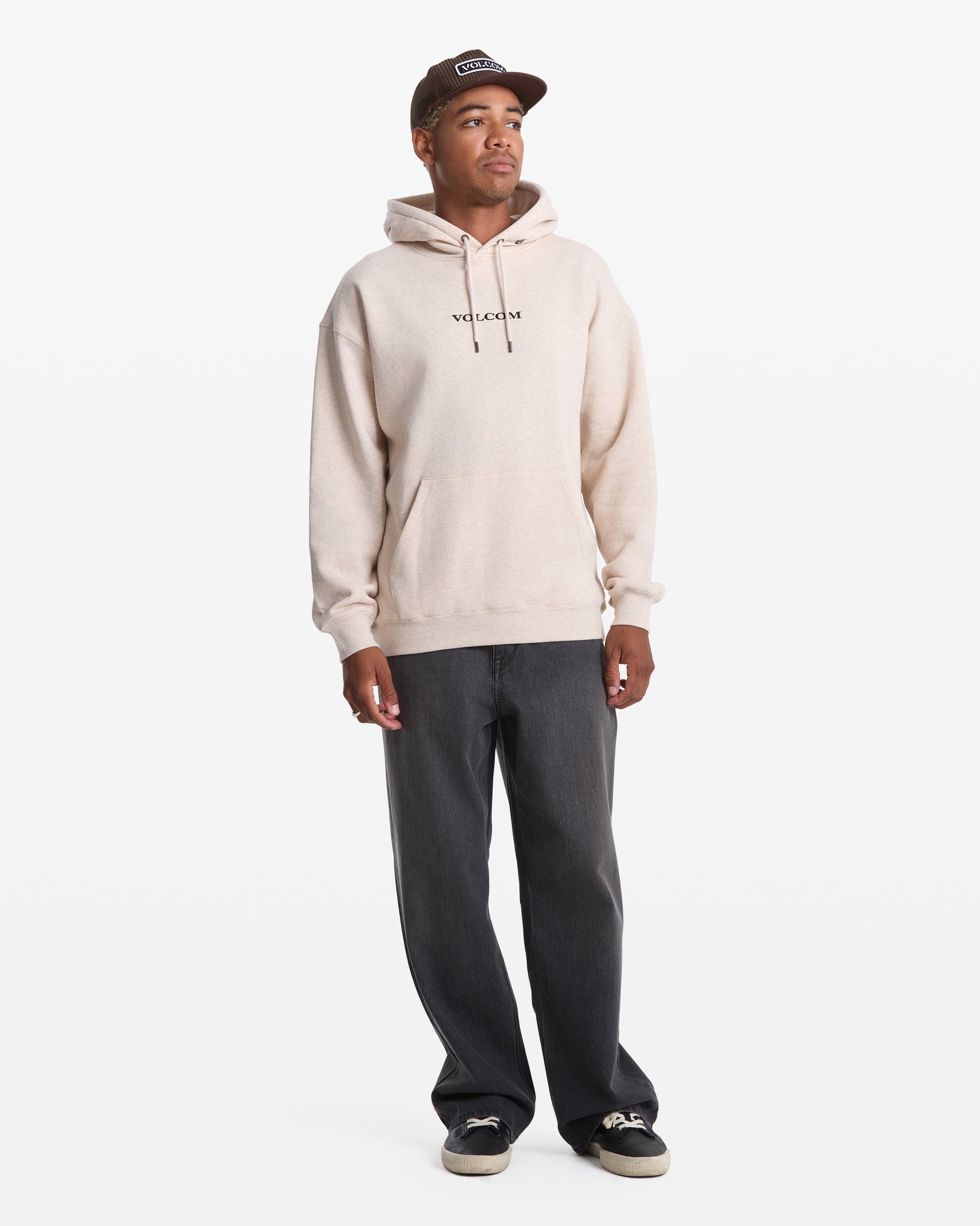 A man wears a beige Men's Volcom Stone Pullover by Volcom, paired with dark gray jeans, black sneakers, and a brown cap as he stands against a plain white background.