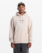 A person wearing a light beige Men's Volcom Stone Pullover by Volcom, dark pants, and a brown cap stands facing forward against a plain white background.