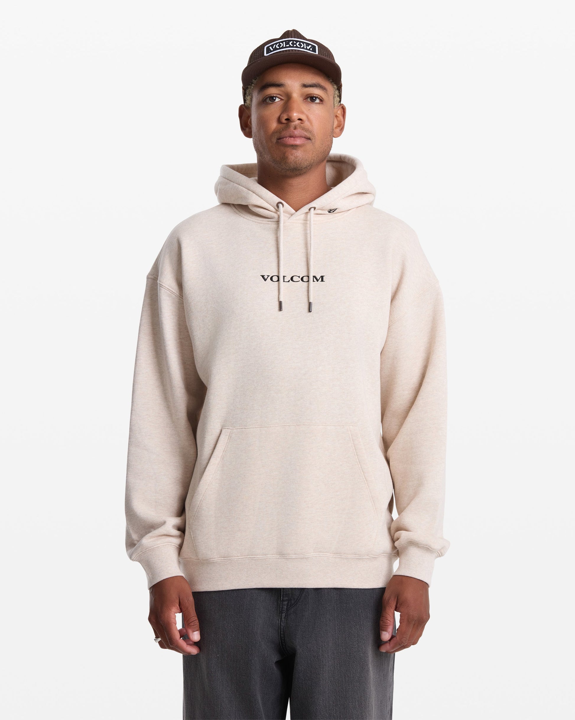 A person wearing a light beige Men's Volcom Stone Pullover by Volcom, dark pants, and a brown cap stands facing forward against a plain white background.
