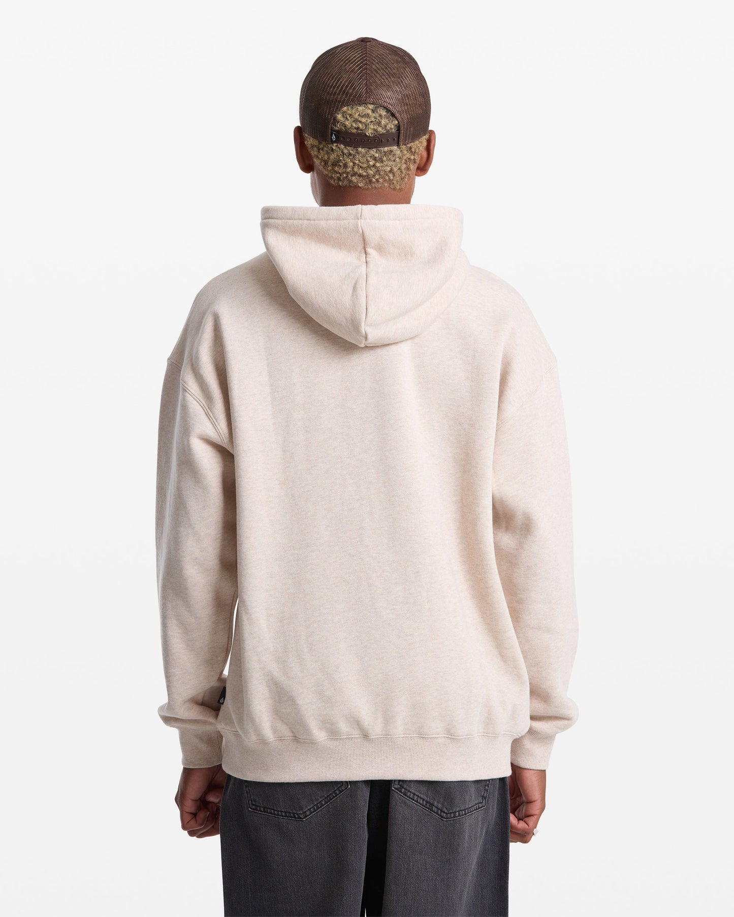A person wearing a light beige Volcom Men's Stone Pullover hoodie and a brown mesh baseball cap stands with their back to the camera against a plain white background.