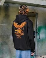 A person with wavy brown hair is seen from behind wearing the Volcom Men's Hammered Pullover, a black hoodie featuring a large orange eagle graphic and VOLCOM Workwear text on the back, standing by a weathered green and gray wall.