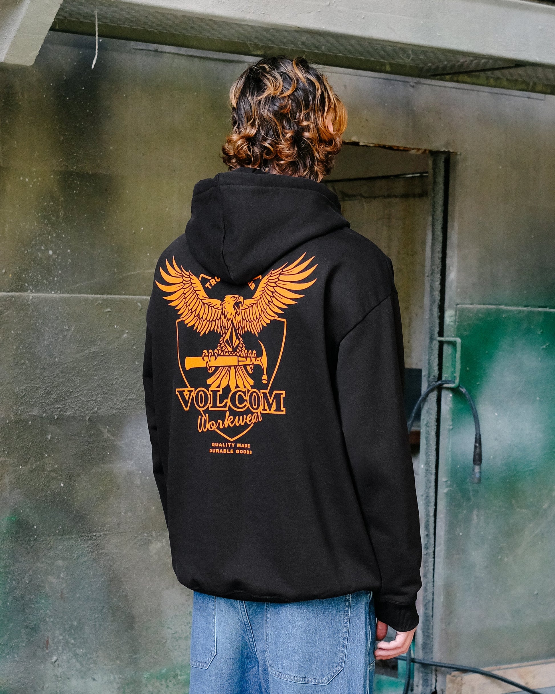 A person with wavy brown hair is seen from behind wearing the Volcom Men's Hammered Pullover, a black hoodie featuring a large orange eagle graphic and VOLCOM Workwear text on the back, standing by a weathered green and gray wall.