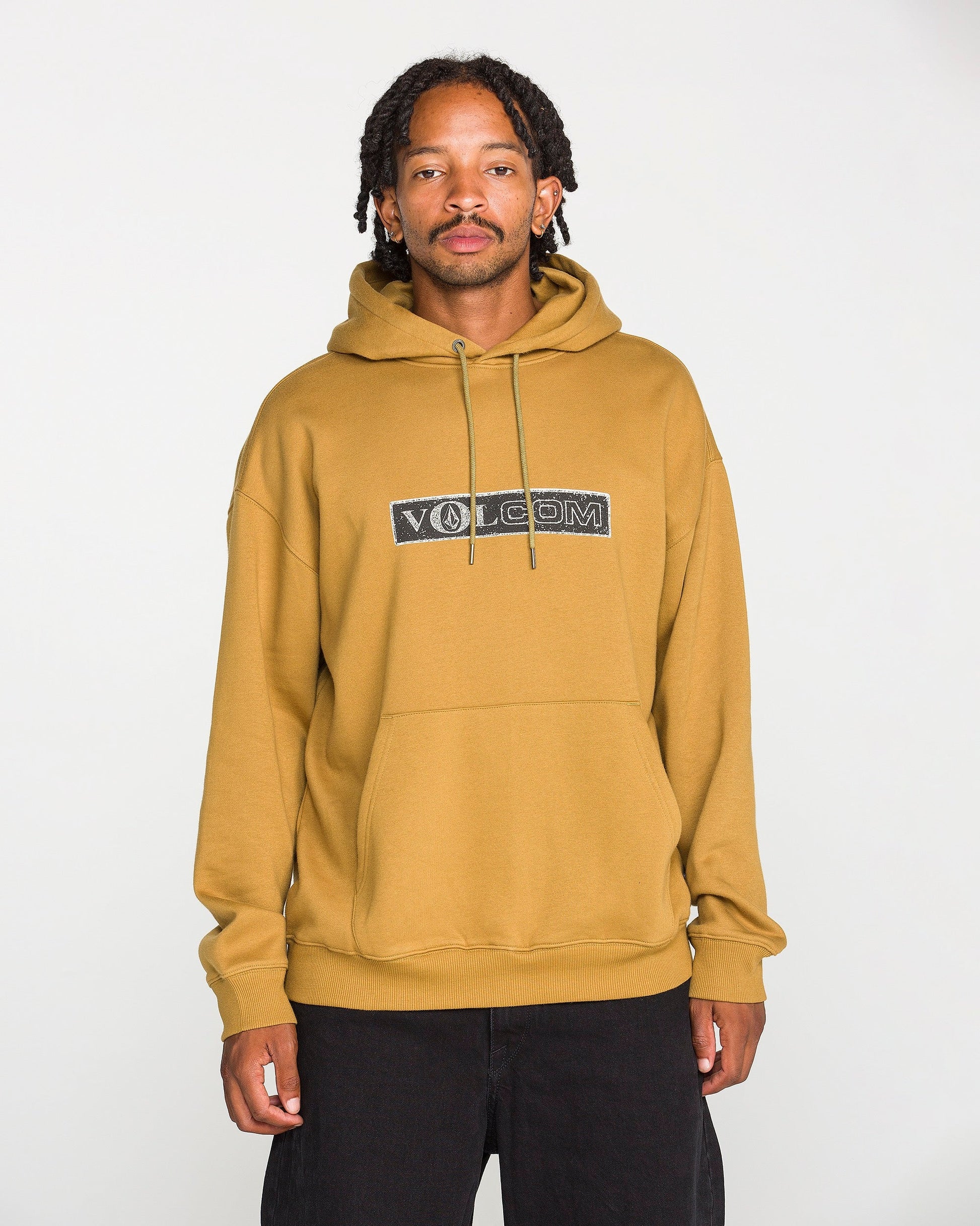A person wearing the Volcom Men's Super Stoned Pullover in mustard yellow, crafted from brushed-back fleece, paired with black pants, stands against a plain white background and looks at the camera.