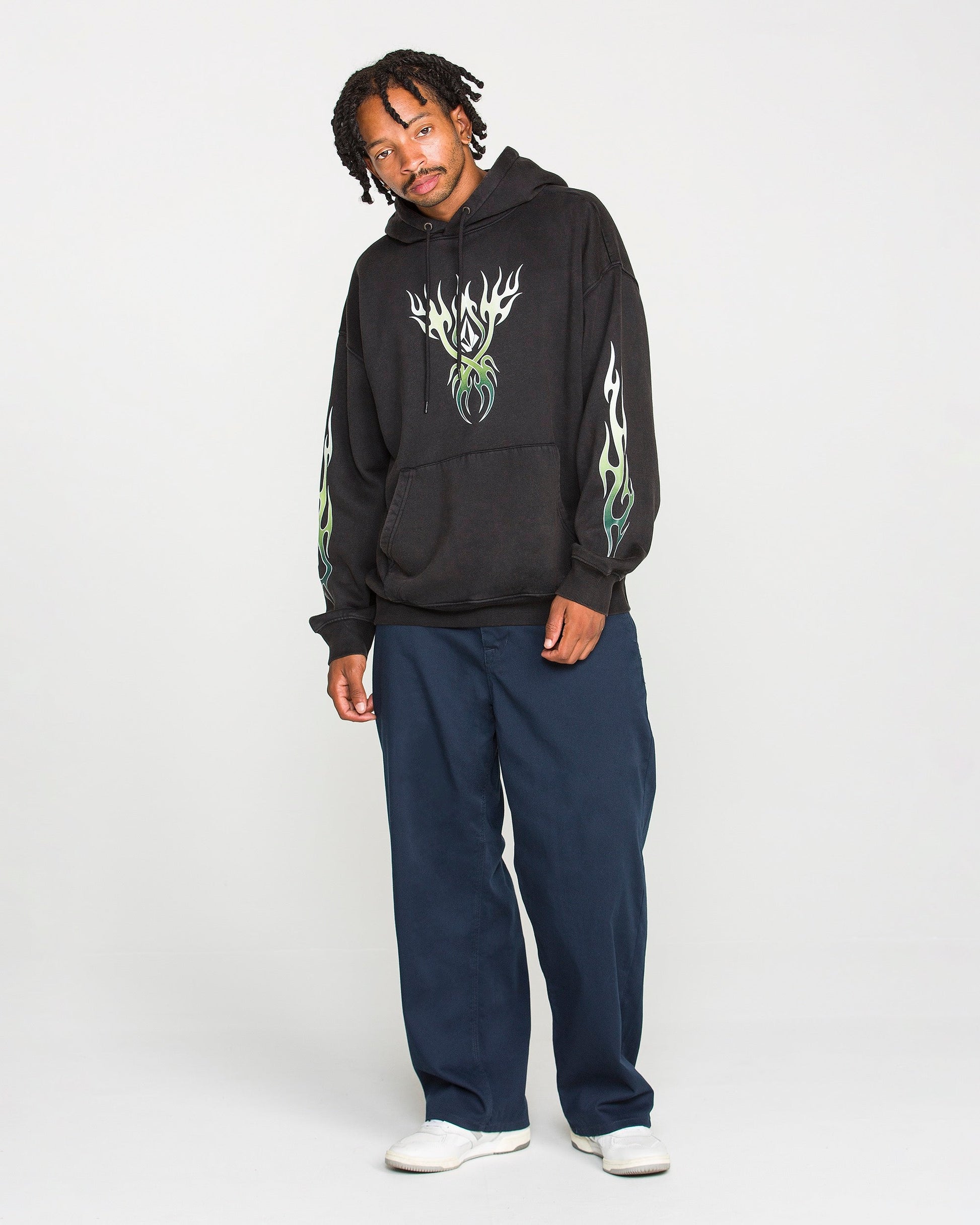 A person stands against a plain background wearing the Volcom Men's Stone Trip Pullover, a washed-black tribal hoodie featuring striking green flame designs. Paired with navy blue pants and white shoes, they pose relaxedly with a slight smile. The heavyweight fleece fabric of the hoodie adds both comfort and style to the overall look.