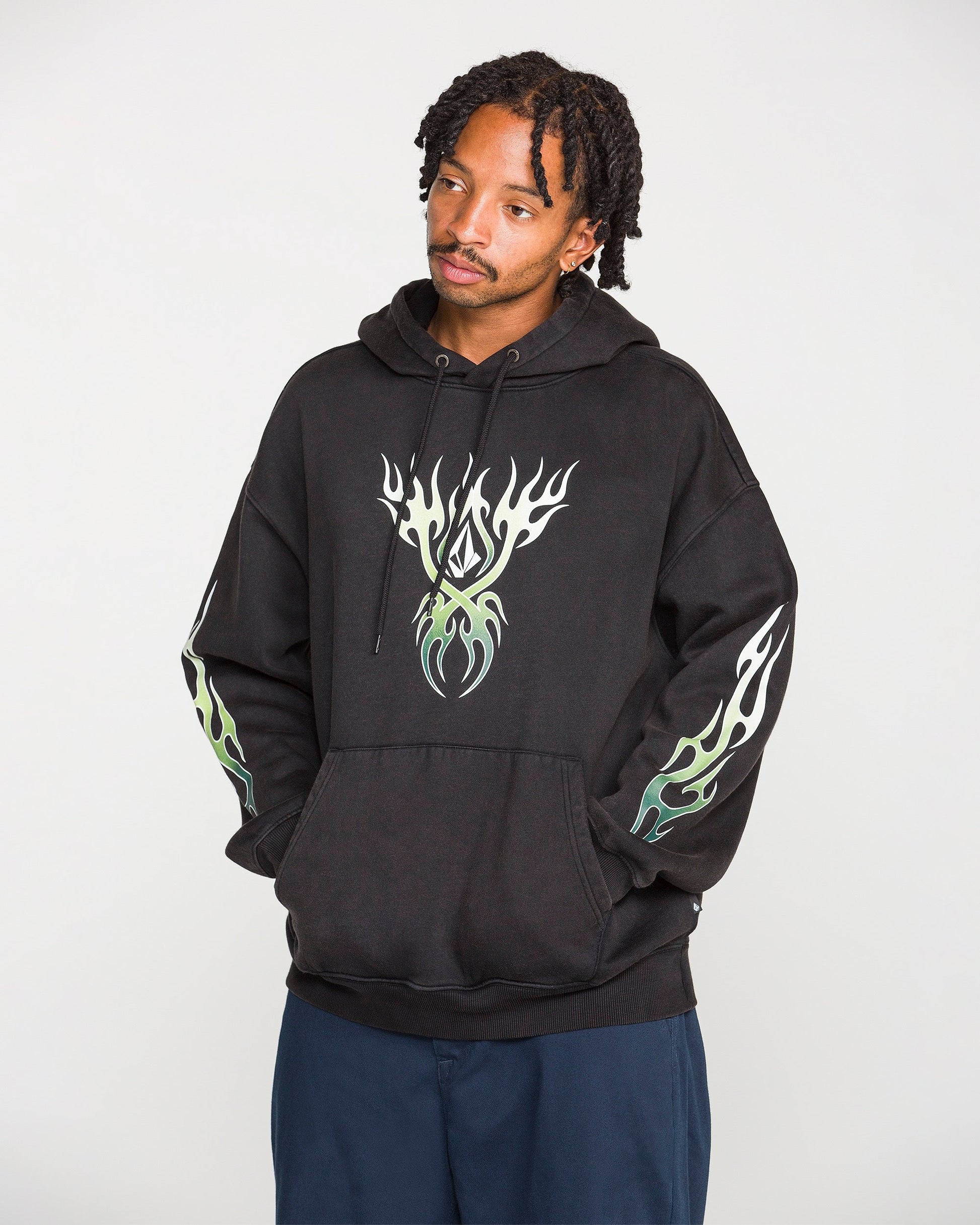 A person wearing the Volcom Men's Stone Trip Pullover—a washed-black hoodie featuring white and green flame designs on the chest and sleeves—stands with hands in the front pocket, looking slightly to the side.