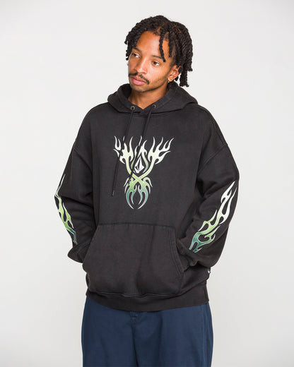 A person wearing the Volcom Men's Stone Trip Pullover—a washed-black hoodie featuring white and green flame designs on the chest and sleeves—stands with hands in the front pocket, looking slightly to the side.