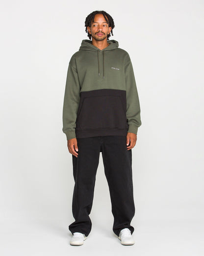 A person stands facing forward, wearing the Volcom Men's Long Division Pullover—a green and black colorblocked hoodie made with REPREVE recycled fibers—paired with black pants and white sneakers, against a plain white background.