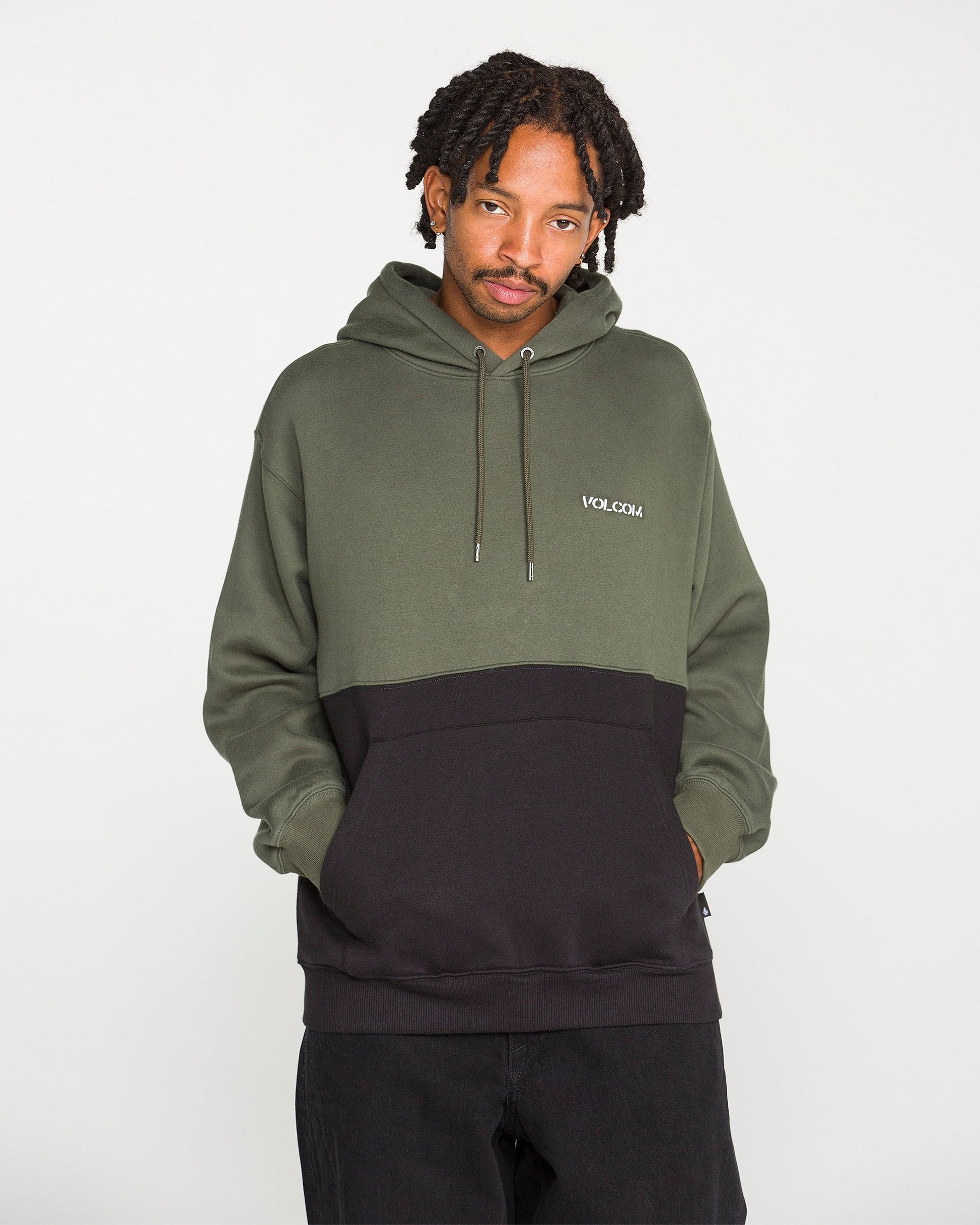 A person stands with hands in pockets, looking at the camera while wearing the Volcom Men's Long Division Pullover—a green and black colorblocked hoodie made with REPREVE recycled fibers and featuring "VOLCOM" on the chest. The background is plain white.