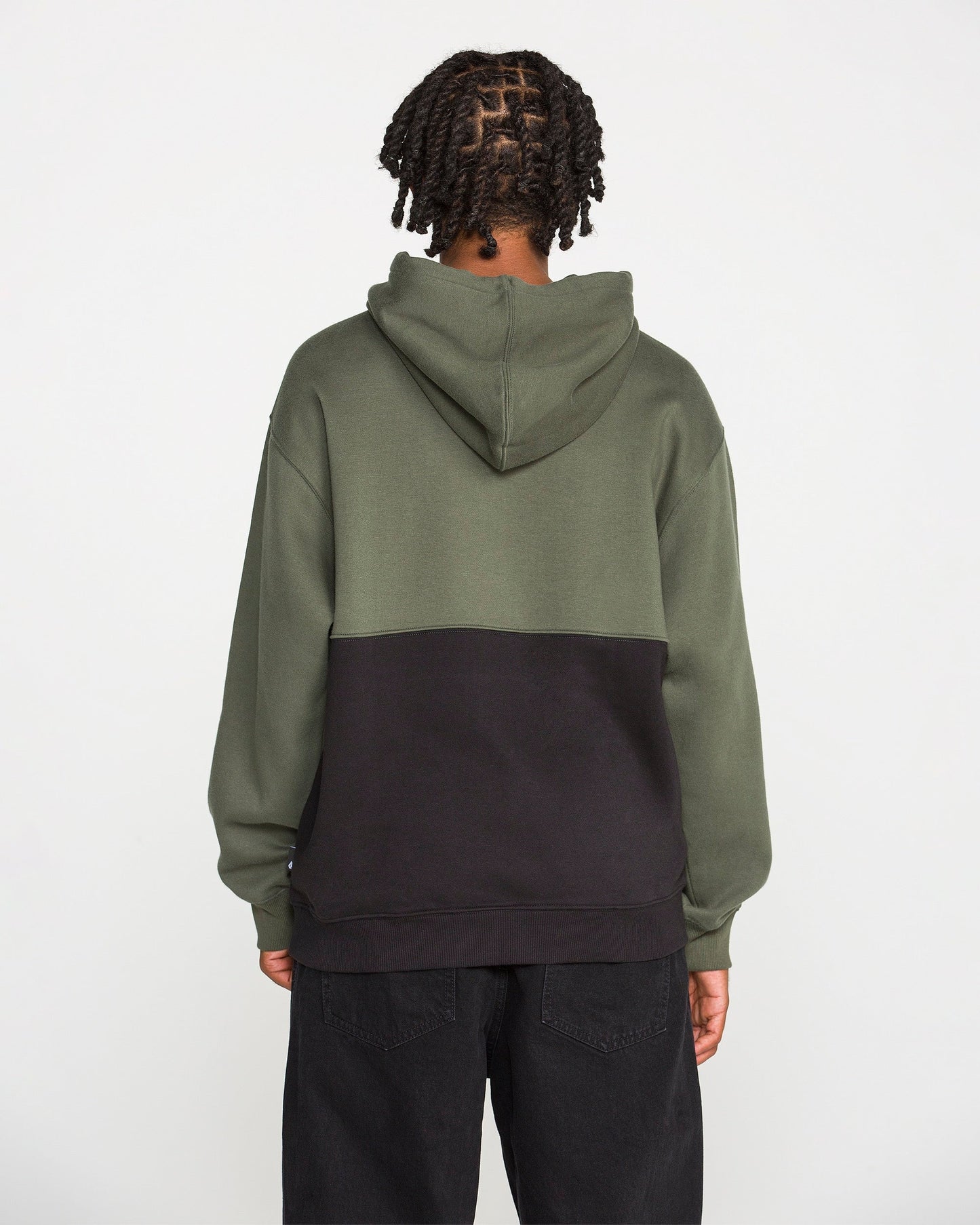 A person with braided hair is seen from behind against a plain white background, wearing the Volcom Men's Long Division Pullover. The hoodie showcases green and black colorblocked panels and is paired with black pants. Crafted with REPREVE recycled fibers, this eco-conscious pullover combines style and sustainability.
