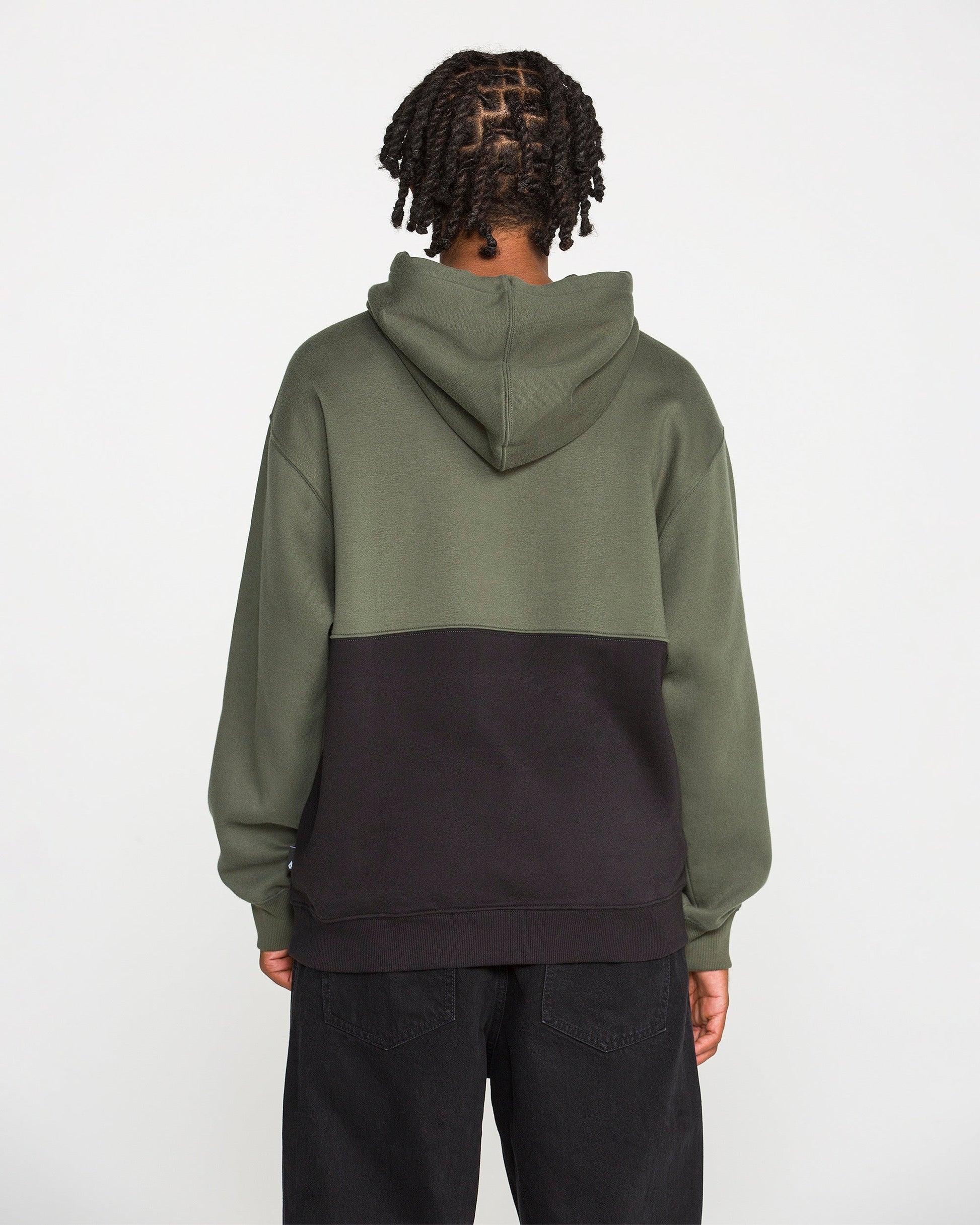 A person with braided hair is seen from behind against a plain white background, wearing the Volcom Men's Long Division Pullover. The hoodie showcases green and black colorblocked panels and is paired with black pants. Crafted with REPREVE recycled fibers, this eco-conscious pullover combines style and sustainability.