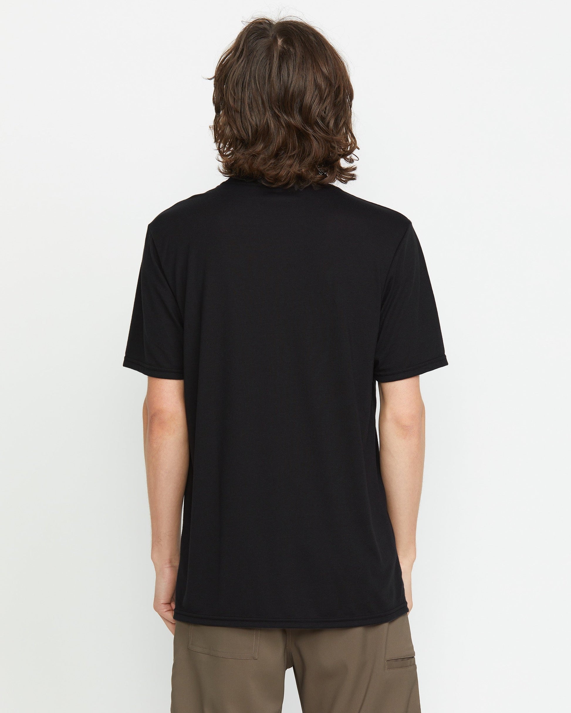 A person with medium-length brown hair is seen from behind, wearing a Volcom Men's Stone Tech Short Sleeve Tee in black and brown pants, standing against a plain white background.