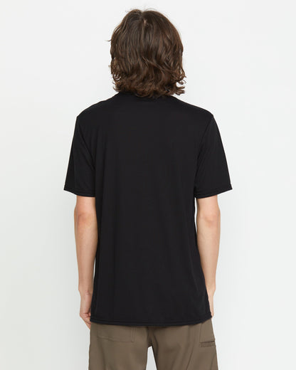 A person with medium-length brown hair is seen from behind, wearing a Volcom Men's Stone Tech Short Sleeve Tee in black and brown pants, standing against a plain white background.