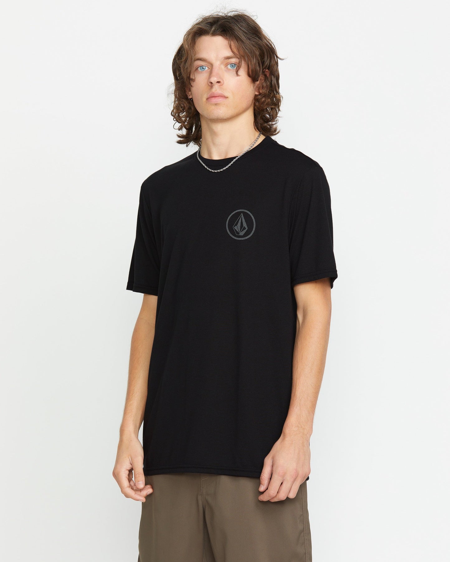 A young man with wavy brown hair wears the Volcom Men's Stone Tech Short Sleeve Tee, a black t-shirt with a reflective round chest logo, paired with khaki pants, standing against a plain white background.