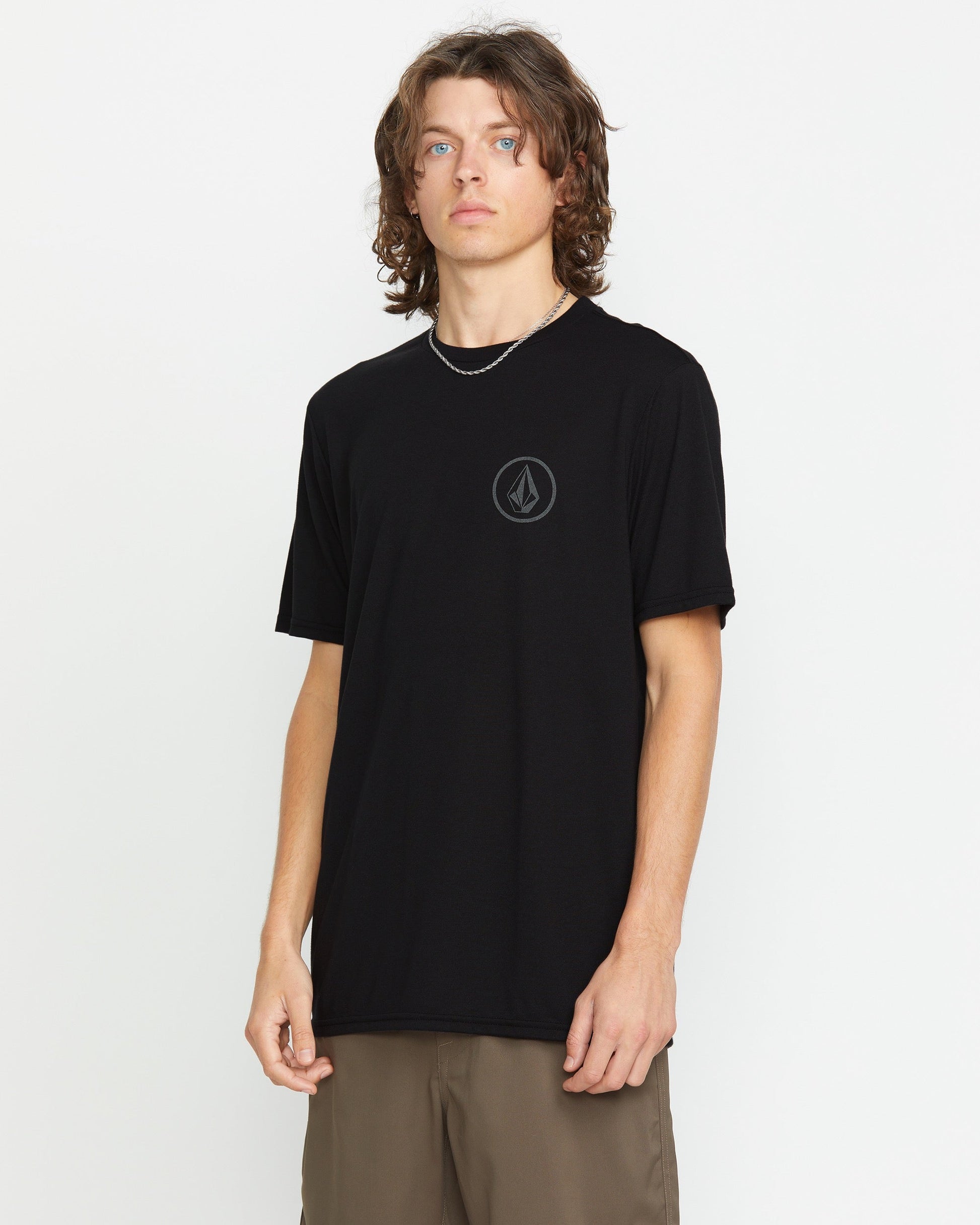 A young man with wavy brown hair wears the Volcom Men's Stone Tech Short Sleeve Tee, a black t-shirt with a reflective round chest logo, paired with khaki pants, standing against a plain white background.