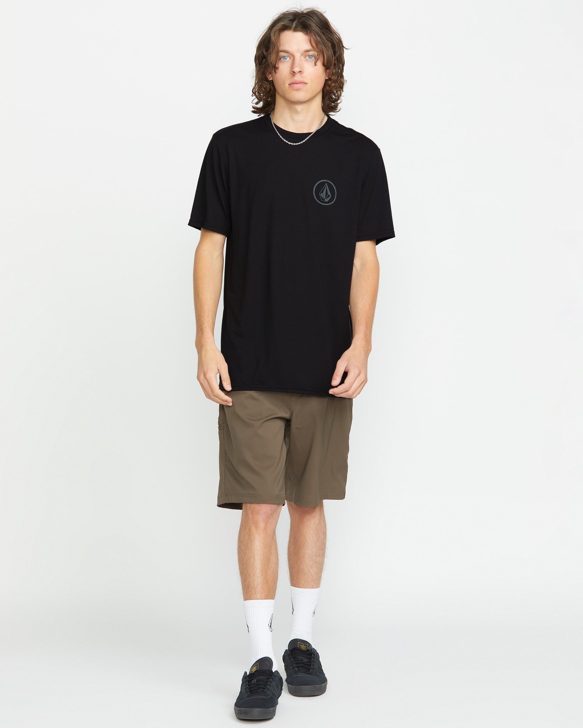 A young man with wavy hair faces the camera, wearing the Volcom Men's Stone Tech Short Sleeve Tee in black, paired with brown shorts, white socks, and black sneakers against a plain white background.