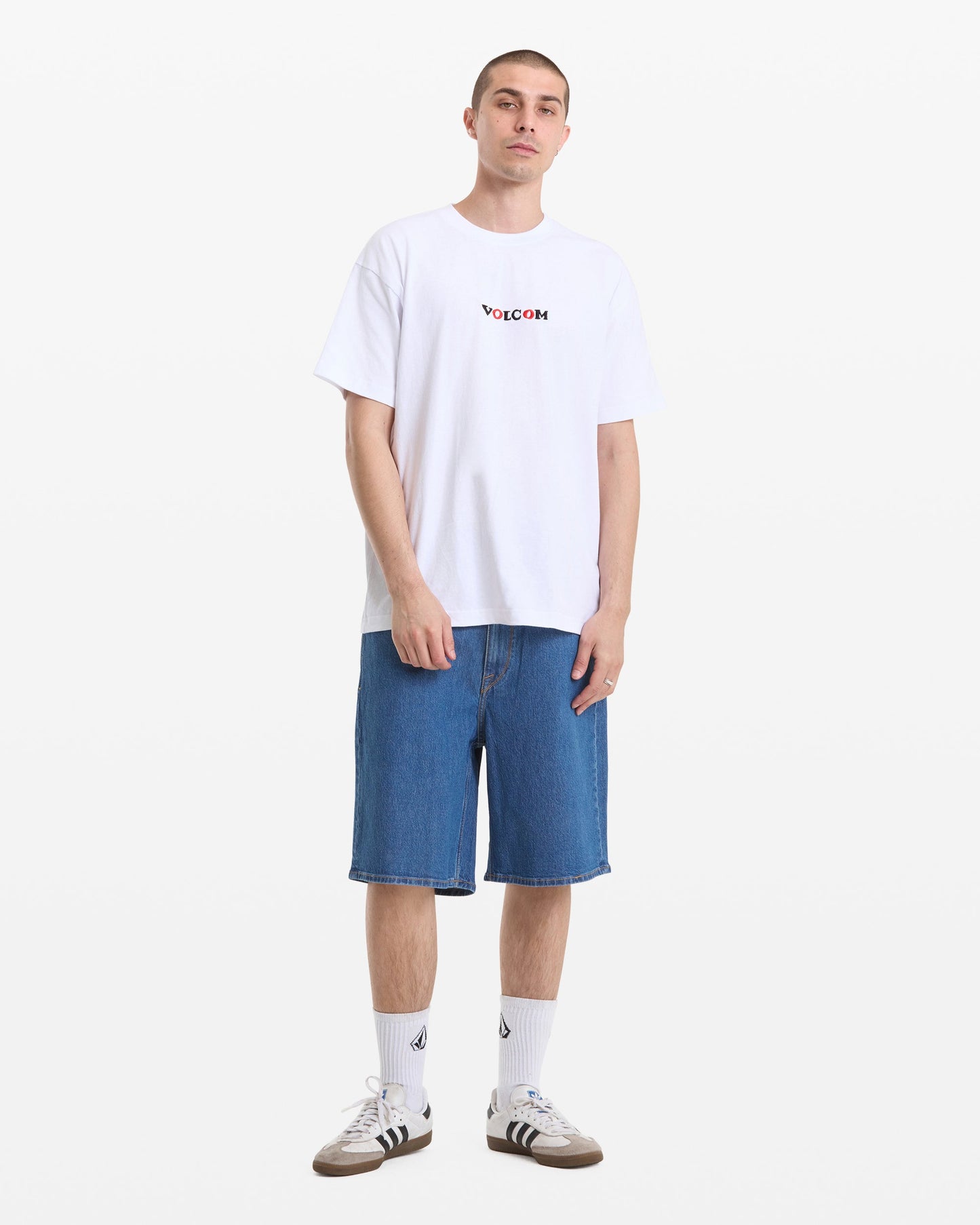 A person stands against a white background wearing a Men's Featured Artist Seth Conboy Loose Short Sleeve Tee by Volcom, knee-length blue denim shorts, white socks with a logo, and gray Adidas sneakers.