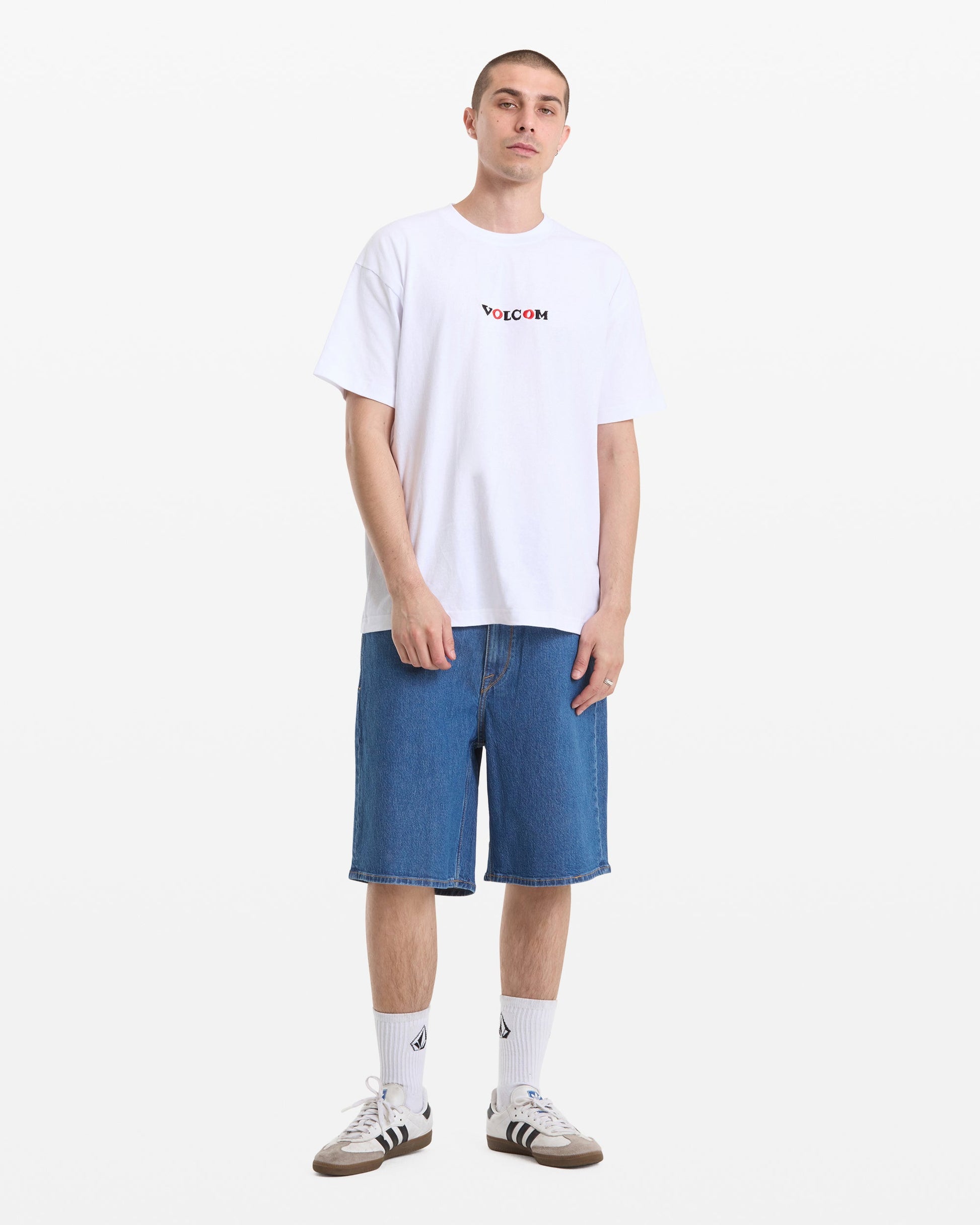 A person stands against a white background wearing a Men's Featured Artist Seth Conboy Loose Short Sleeve Tee by Volcom, knee-length blue denim shorts, white socks with a logo, and gray Adidas sneakers.