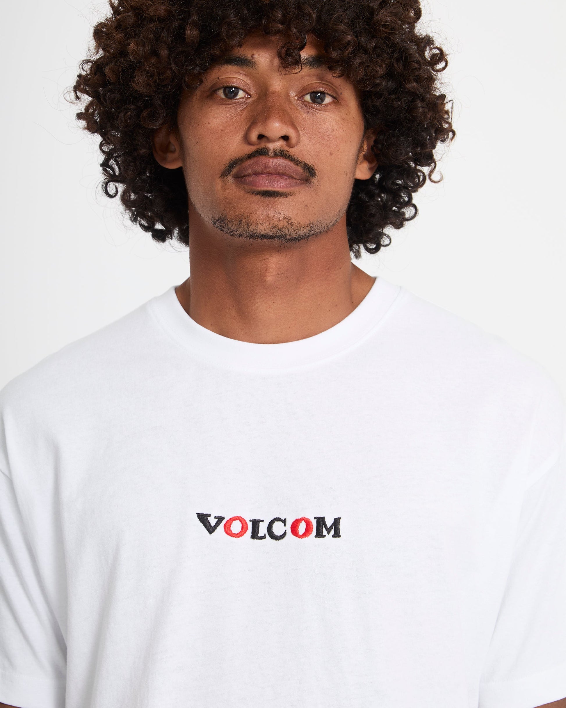 A man with curly hair and a mustache wears the Volcom Men's Featured Artist Seth Conboy Loose Short Sleeve Tee, featuring black and red VOLCOM lettering, standing against a plain white background.