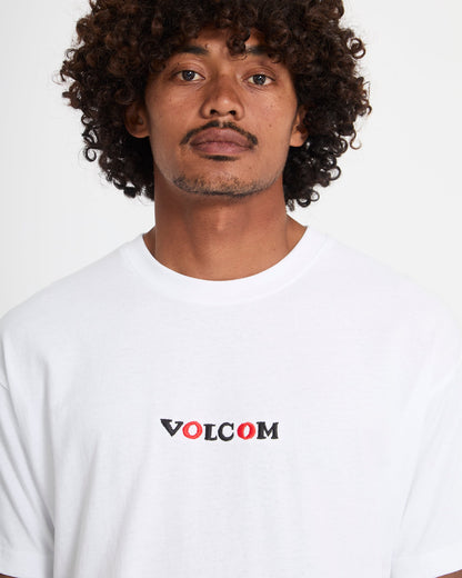 A man with curly hair and a mustache wears the Volcom Men's Featured Artist Seth Conboy Loose Short Sleeve Tee, featuring black and red VOLCOM lettering, standing against a plain white background.