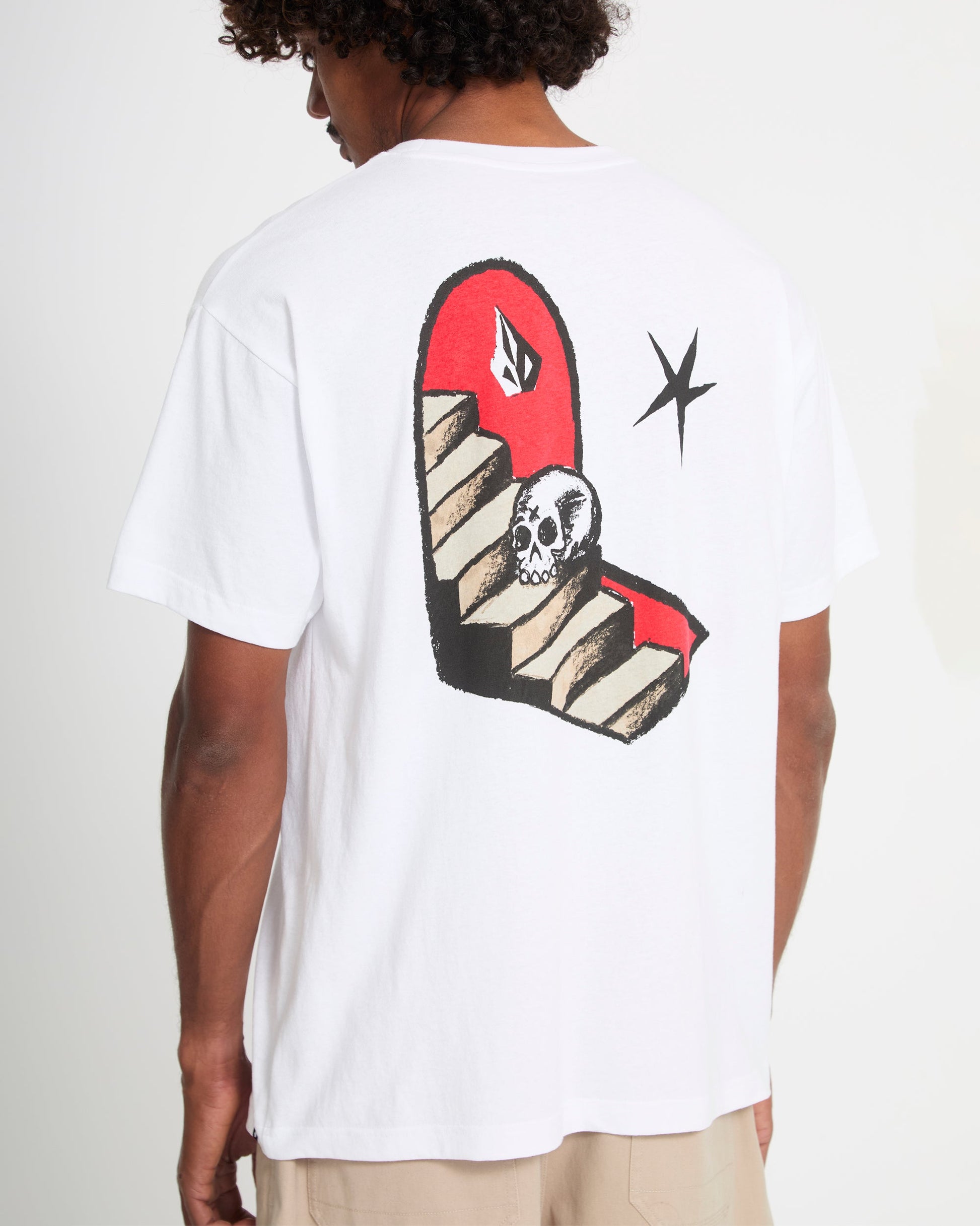 A person is wearing the Volcom Men's Featured Artist Seth Conboy Loose Short Sleeve Tee, a white shirt featuring a back graphic of a red staircase, skull at the bottom, black star, and abstract designs near the top.
