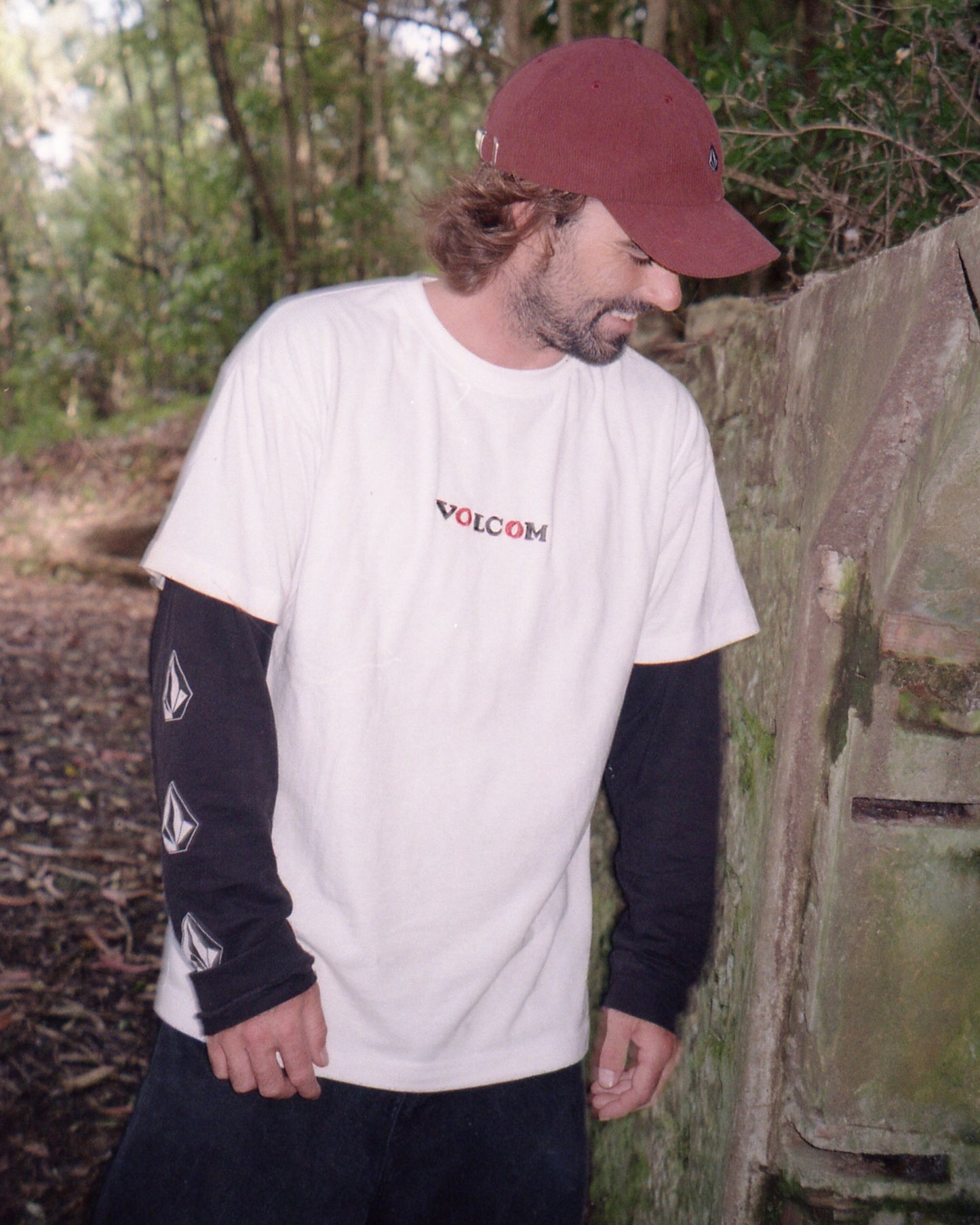 A bearded man in a red cap stands outdoors near a mossy wall, wearing a white Volcom Men's Featured Artist Seth Conboy Loose Short Sleeve Tee over a black long-sleeve shirt in a wooded area.