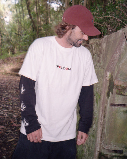 A bearded man in a red cap stands outdoors near a mossy wall, wearing a white Volcom Men's Featured Artist Seth Conboy Loose Short Sleeve Tee over a black long-sleeve shirt in a wooded area.