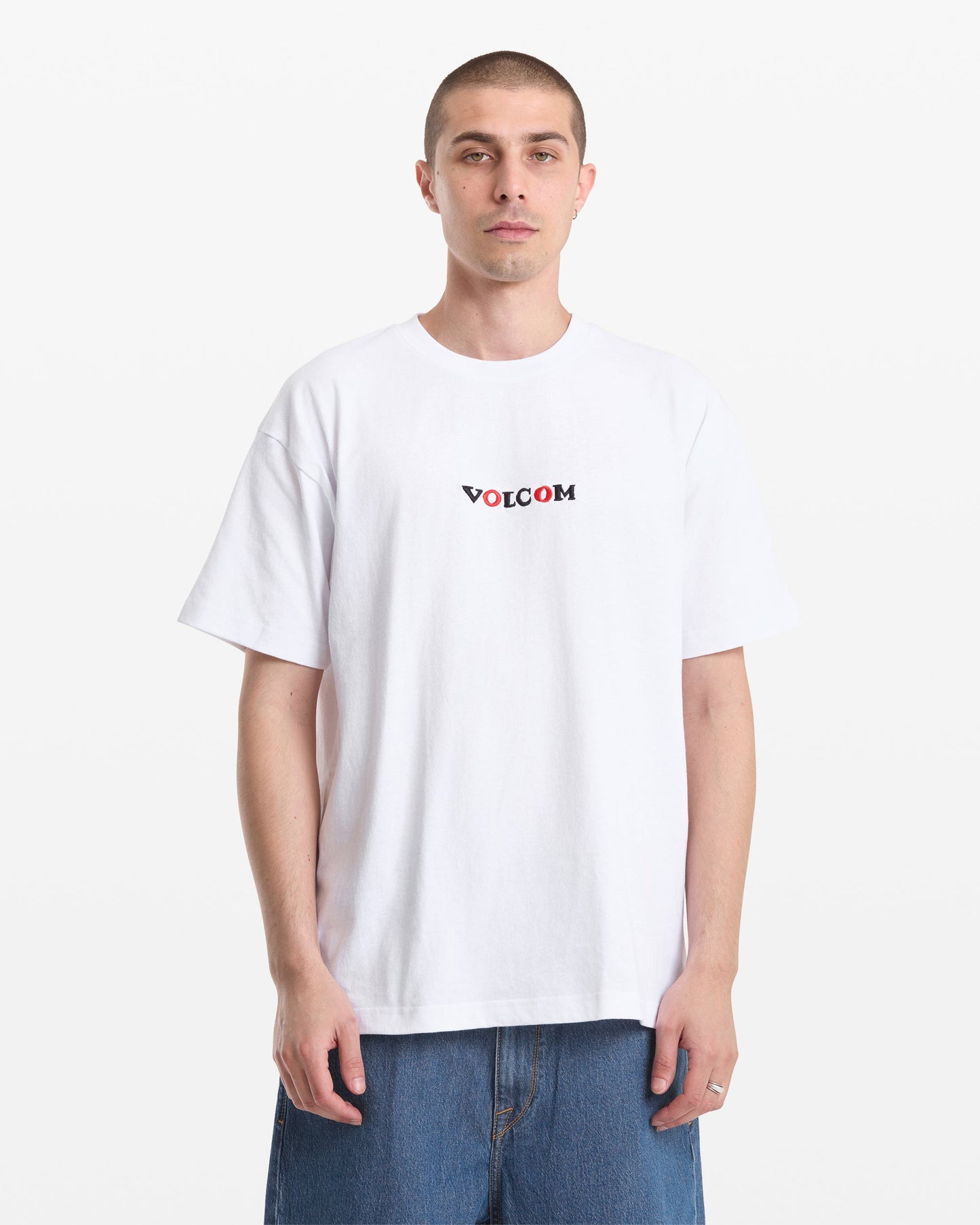 A man with short hair wears the Volcom Men's Featured Artist Seth Conboy Loose Short Sleeve Tee and blue denim shorts, standing against a plain white background.