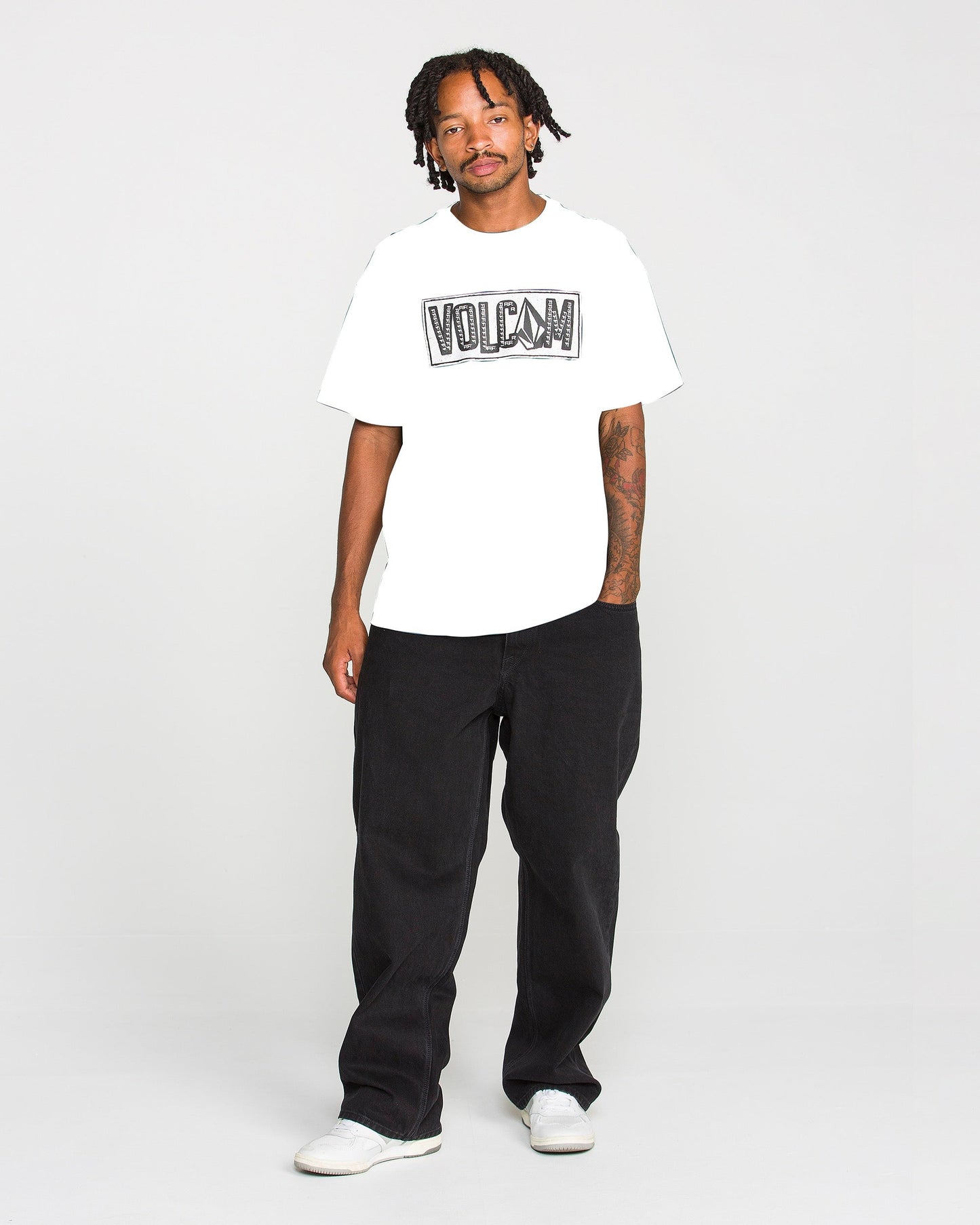 A person stands against a plain background, wearing the Volcom Men's Studded Short Sleeve Tee in white, crafted from heavyweight premium cotton, paired with black pants and white shoes. One arm displays a tattoo, while the tee features a soft-hand screen print that brings subtle style to its classic design.